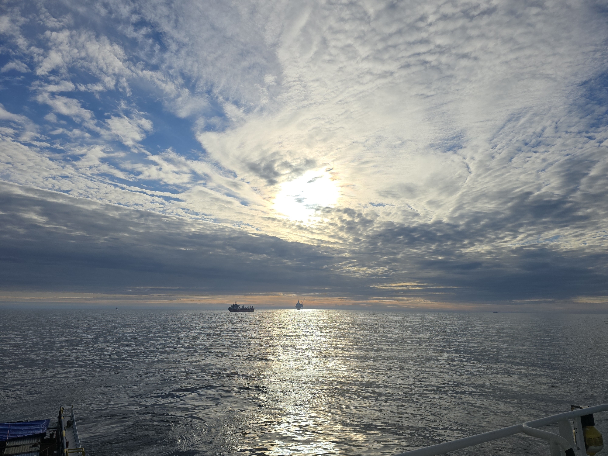 The image shows a seascape under a sky with patchy clouds obscuring the sun. The water surface is reflective, with a glinting path of light extending from the horizon towards the viewer. Two ships are visible on the horizon, silhouetted against the bright sky. The location is Unknown, Unknown.