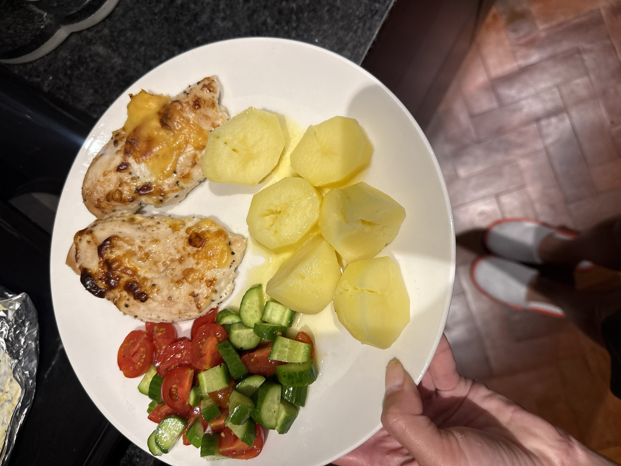 A high-angle shot shows a white plate held by a person. The plate holds a meal consisting of two pieces of what appears to be chicken, some boiled potatoes, and a mixed salad of diced tomatoes and cucumbers. The chicken pieces appear to be topped with melted cheese. The environment is a kitchen with a dark countertop. A person's feet wearing white slippers with a red stripe are visible in the background. There is no indication of the image being taken in Cape Town, South Africa.