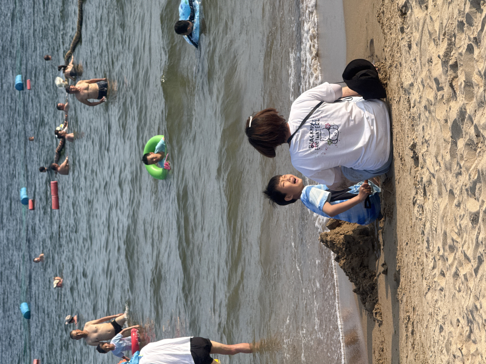 A group of people are enjoying a sunny day at the beach in Qinhuangdao, China, with some swimming in the ocean and others relaxing on the sand. In the foreground, a woman sits on the sand with her back to the camera, wearing a white t-shirt and blue jeans. Beside her, a young boy in a blue and white striped shirt is playing with a pile of sand, his mouth open as if laughing. In the background, several people are in the water, some with inflatable rings, and a long, brown rope snakes across the surface of the sea. The water is calm with gentle waves lapping the shore. The sand is light brown and appears well-trodden.