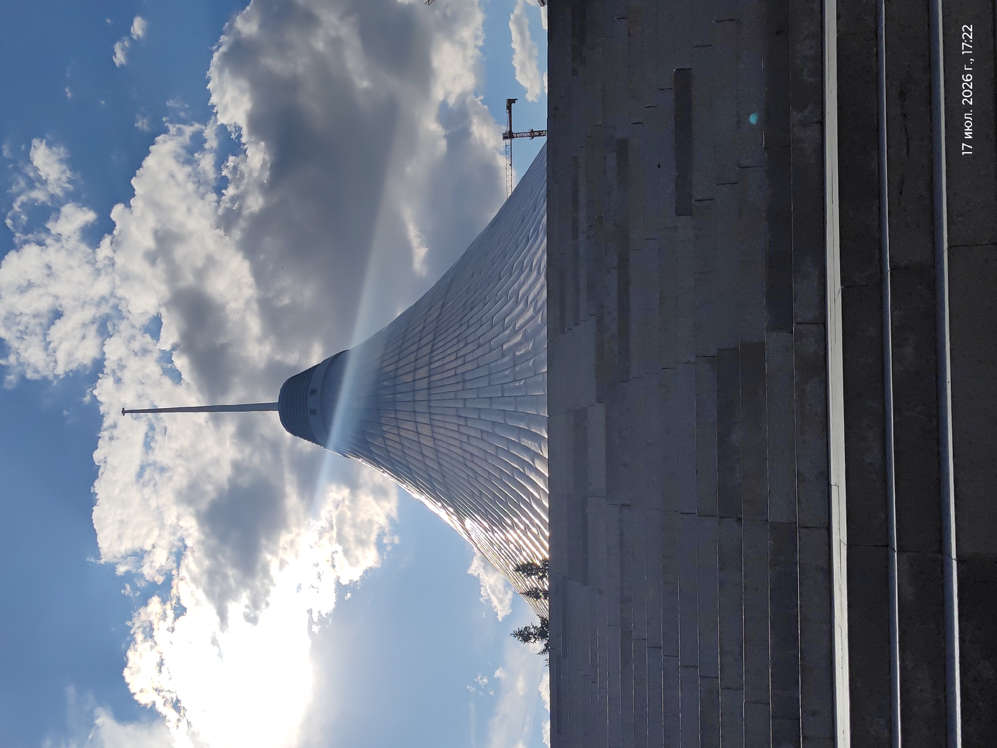 The image shows the Khan Shatyr Entertainment Center in Astana, Kazakhstan, a distinctive tent-shaped building with a tall spire. The structure is covered in a metallic, scale-like material that reflects the sunlight. In the foreground, a wide, grey stone staircase leads upwards towards the building. The sky above is blue with scattered white and grey clouds, and the sun is visible, casting a bright glare. To the right of the building, a construction crane is partially visible against the sky. The bottom right corner of the image contains a timestamp: "17 июл. 2026 г., 17:22". There are no people visible in the image. The overall scene depicts a large architectural landmark under a dynamic sky, with evidence of ongoing construction in the background.