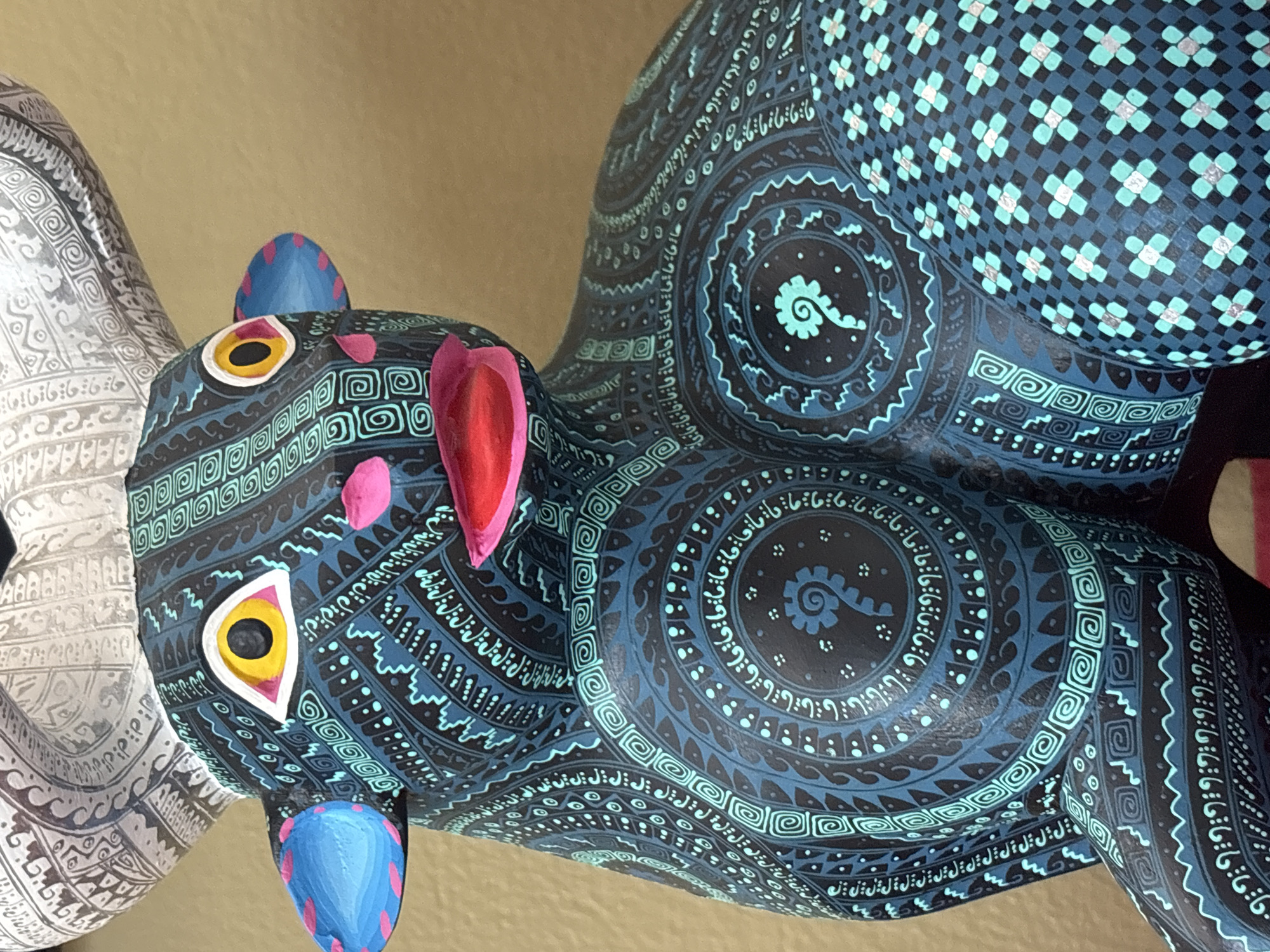 The image displays a close-up view of a vibrantly painted Alebrije. The Alebrije is adorned with intricate, geometric patterns in shades of blue, black, and turquoise. Other colors like red, pink, yellow, and white are also noticeable, particularly around the face and ears. The Alebrije is likely an animal figure, given its apparent shape.