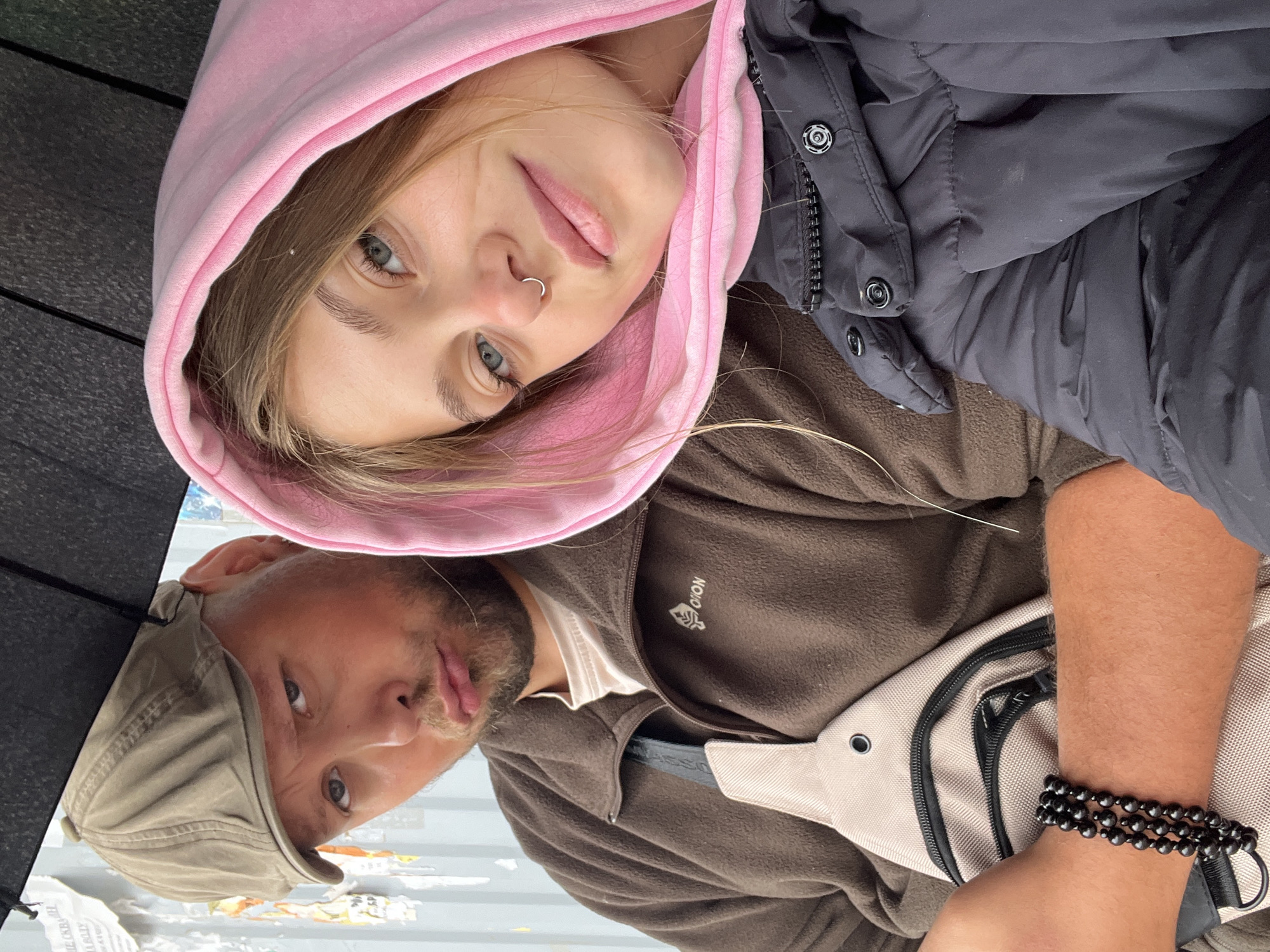 Two people are captured in a selfie-style shot. The person on top is a young woman with light hair, wearing a pink hooded sweatshirt and a black jacket. She has fair skin and a nose ring. Below her is a man wearing a brown fleece jacket, a khaki cap, and a white t-shirt. He has a light-colored beard. A black bracelet adorns his wrist, which rests on a beige-colored fanny pack. An open black umbrella is visible in the top-left of the frame. The background shows an urban setting with posters and other debris, suggesting a street scene in a city. The location is unspecified; the inclusion of "Фоки, Russia" in the query does not inherently indicate the photo was taken there.