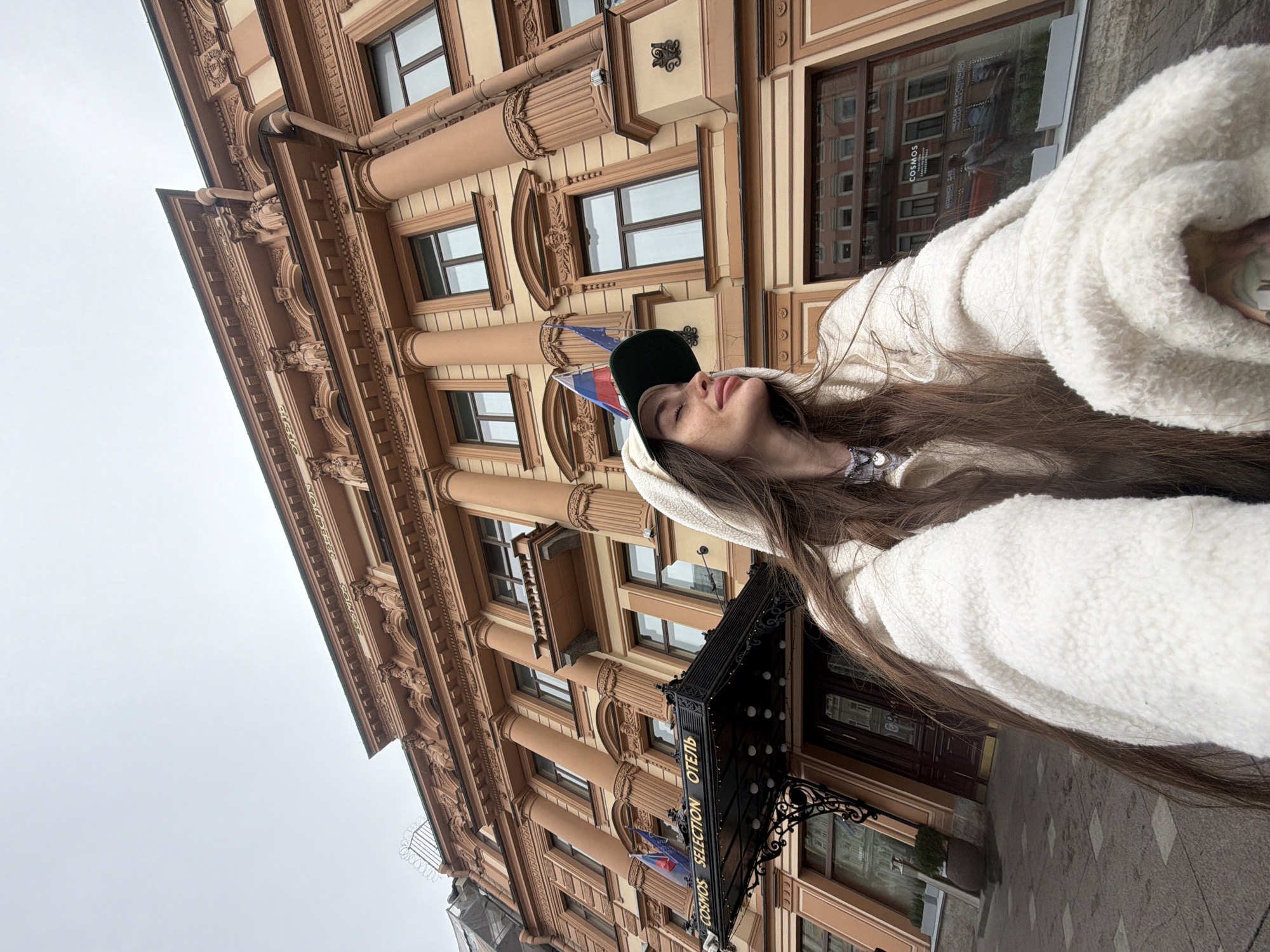 A young woman, with long brown hair and a white hooded jacket, stands in front of the Cosmos Selection hotel in Saint Petersburg, Russia. She is wearing a green baseball cap and has her eyes closed, tilting her head back. The hotel is a large, ornate building with columns and decorative stonework. Two flags, one Russian and one possibly European, hang above the hotel entrance. The sky is overcast.
