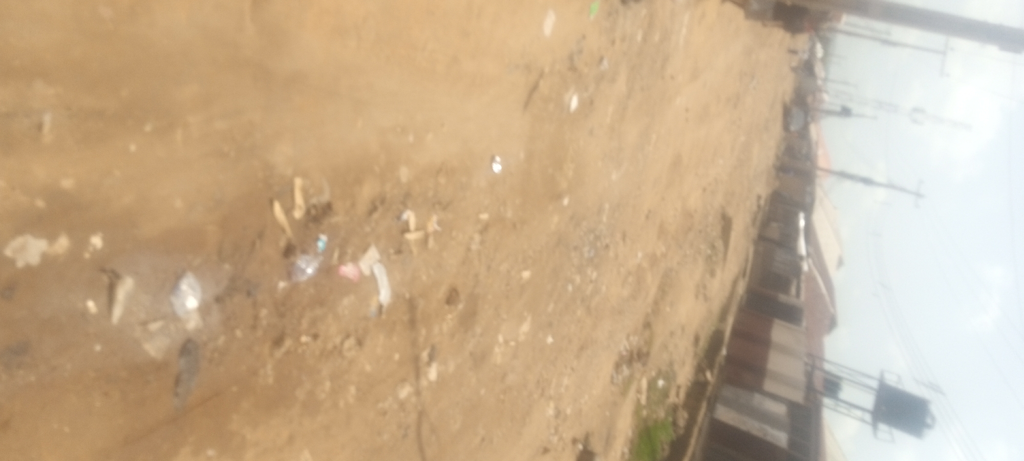 The image depicts a dirt road in Narayi, Nigeria, littered with trash. Buildings are visible on one side of the road, with utility poles and wires overhead. No people are visible.