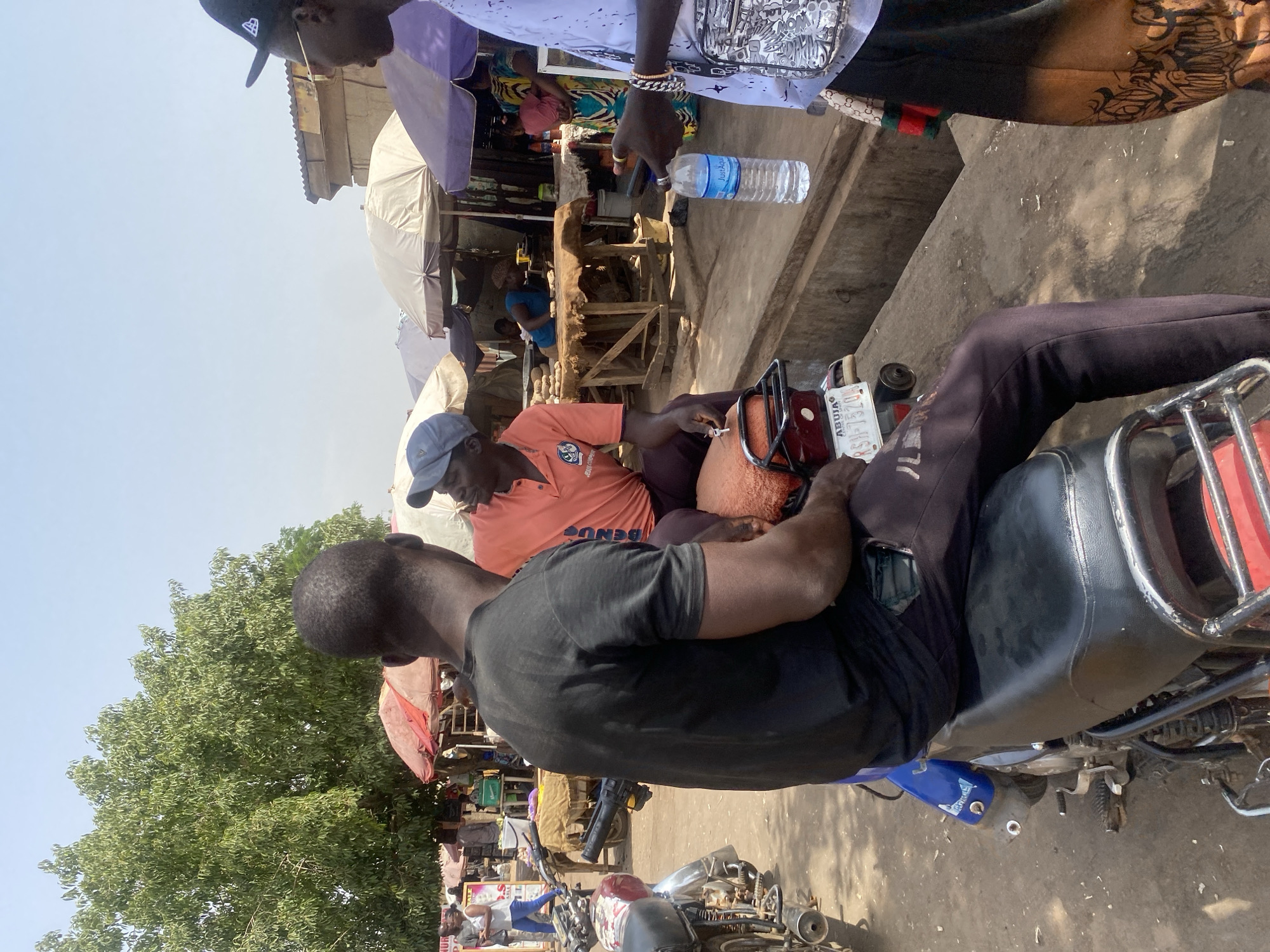 The image portrays an outdoor market scene in Sabon Tasha, Nigeria, under clear daylight conditions. Three adult males are prominent in the foreground. A male figure, seen from the back, is seated on a black motorcycle with a red tail light and a visible license plate displaying "ABUJA RSH-732DS". He wears a black short-sleeved shirt and dark trousers. To his right, another male is seated, facing the first, wearing an orange polo shirt and a light blue baseball cap. A third male stands on the right, facing the two seated individuals, wearing a white short-sleeved top, a black baseball cap, and sunglasses. This standing individual holds a clear plastic bottle labeled "JustAqua" in his left hand and a smartphone in his right hand. A patterned satchel hangs from his right shoulder.
The background features an active market environment with multiple makeshift stalls shaded by various colored umbrellas, including red, beige, and purple. Goods, specifically tubers such as yams or potatoes, are displayed on wooden tables. Additional individuals, consistent with vendors and patrons, are visible in the mid-ground and background. Other motorcycles are present on the unpaved and partially paved ground. A large tree with dense green foliage occupies the upper left background, set against a bright, cloudless sky. Structural elements, including buildings with corrugated metal roofs and concrete walls, are visible behind the market stalls.