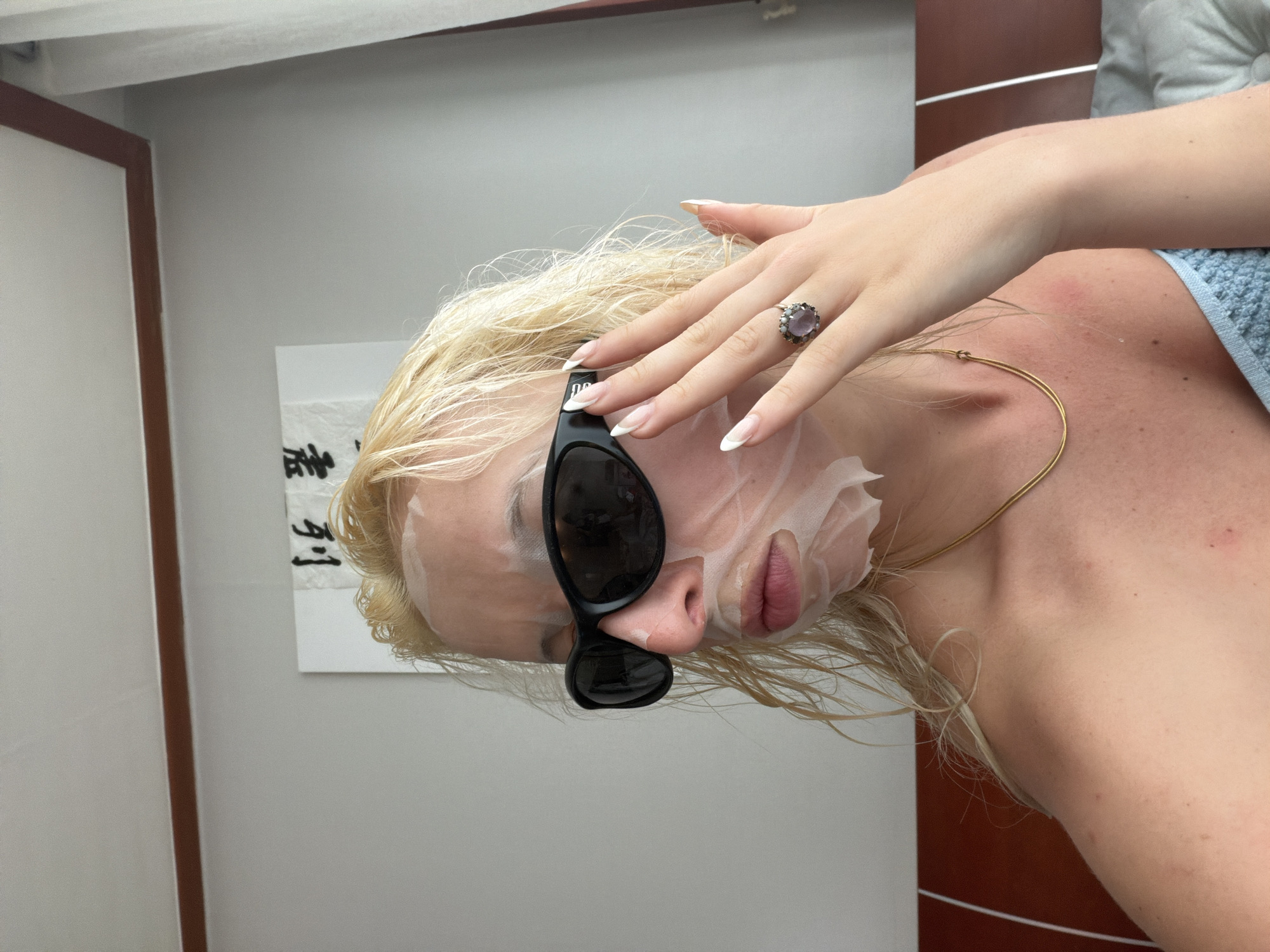 A fair-skinned person with blonde, wet hair is wearing a sheet face mask and black sunglasses. Their left hand is raised towards their face, revealing a ring with a large stone. They are also wearing a thin gold necklace. The background shows a white wall with a framed print featuring East Asian calligraphy, and a partially visible brown panel. The presence of East Asian calligraphy suggests the location might be Seoul, South Korea.