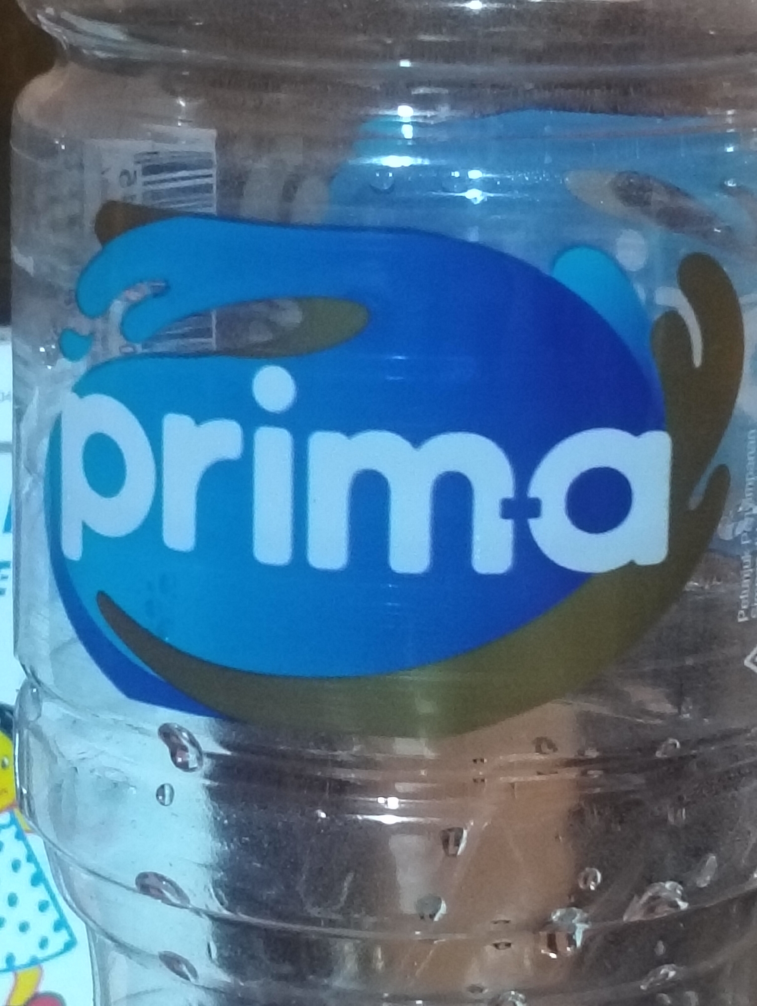 This is a close-up, indoor shot of a clear plastic water bottle with a blue and green label that reads "prima" in white letters. The bottle appears to be full of water, with visible droplets on the exterior. The background is blurry, but it suggests an indoor setting with some lighting. The image does not contain information related to the city of Payagaboh, Indonesia.