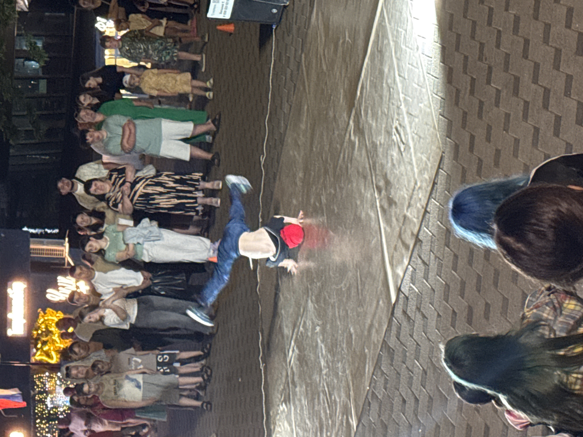 In Unknown, Unknown, a group of people are gathered outdoors at night, watching a breakdancer perform. The breakdancer, wearing blue jeans and a black shirt with a red hat, is in mid-air performing a move, with their body upside down and supported by their hands. The performance is taking place on a large, flat, light-colored surface, possibly a stage or tarp, which is laid out on a cobblestone ground. A speaker is visible to the right of the performer. In the background, other individuals are observing the performance. Some are standing while others are seated, with their backs to the camera, in the foreground. The scene appears to be in an urban area, with some decorative lighting and building facades visible in the background.