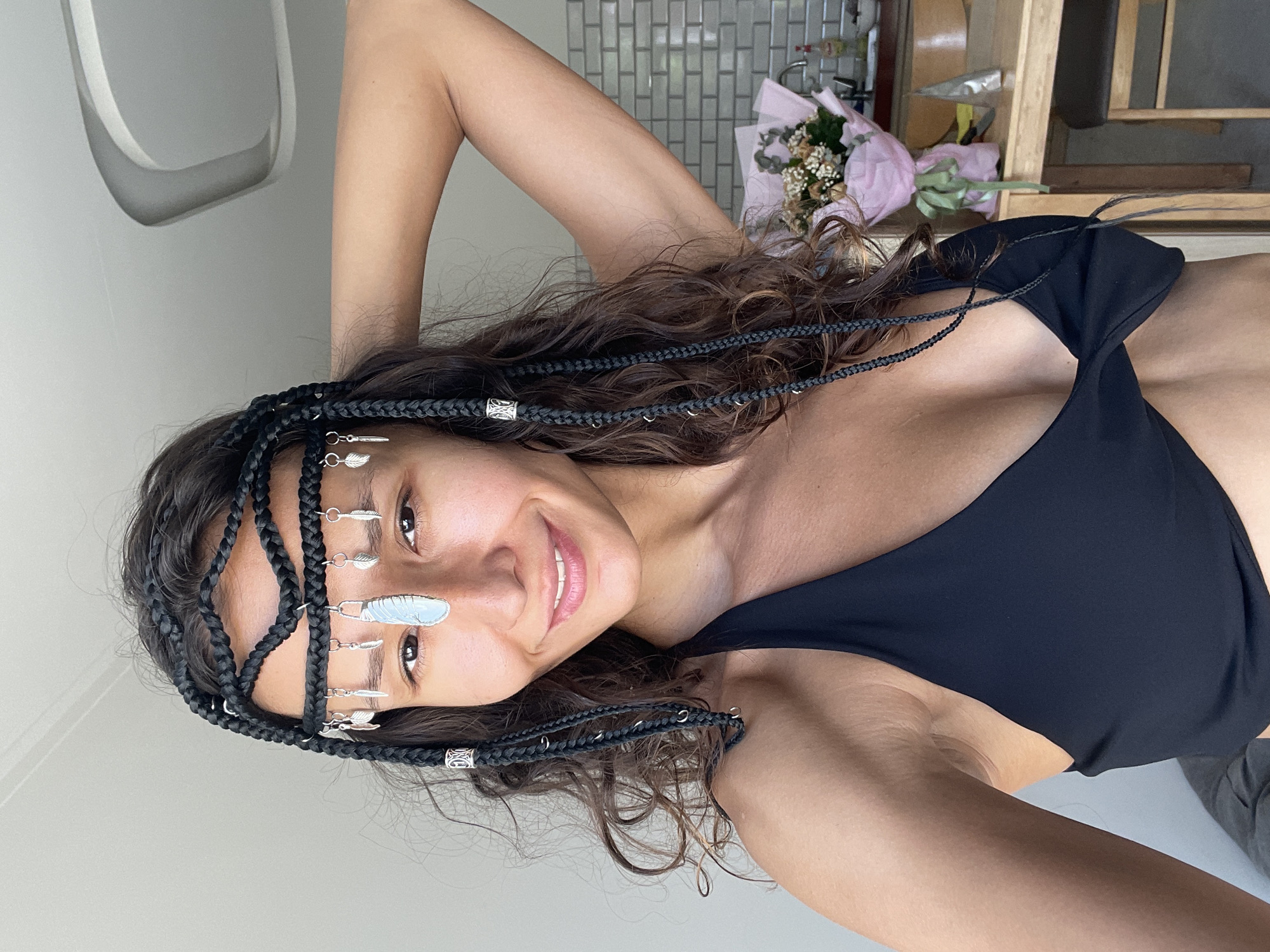 A woman with braided hair and a headpiece with dangling ornaments is shown in a close-up shot. She is wearing a black halter top. The background includes a kitchen counter with a bouquet of flowers and some kitchen appliances. The location is not specified as Mũi Né, Vietnam.