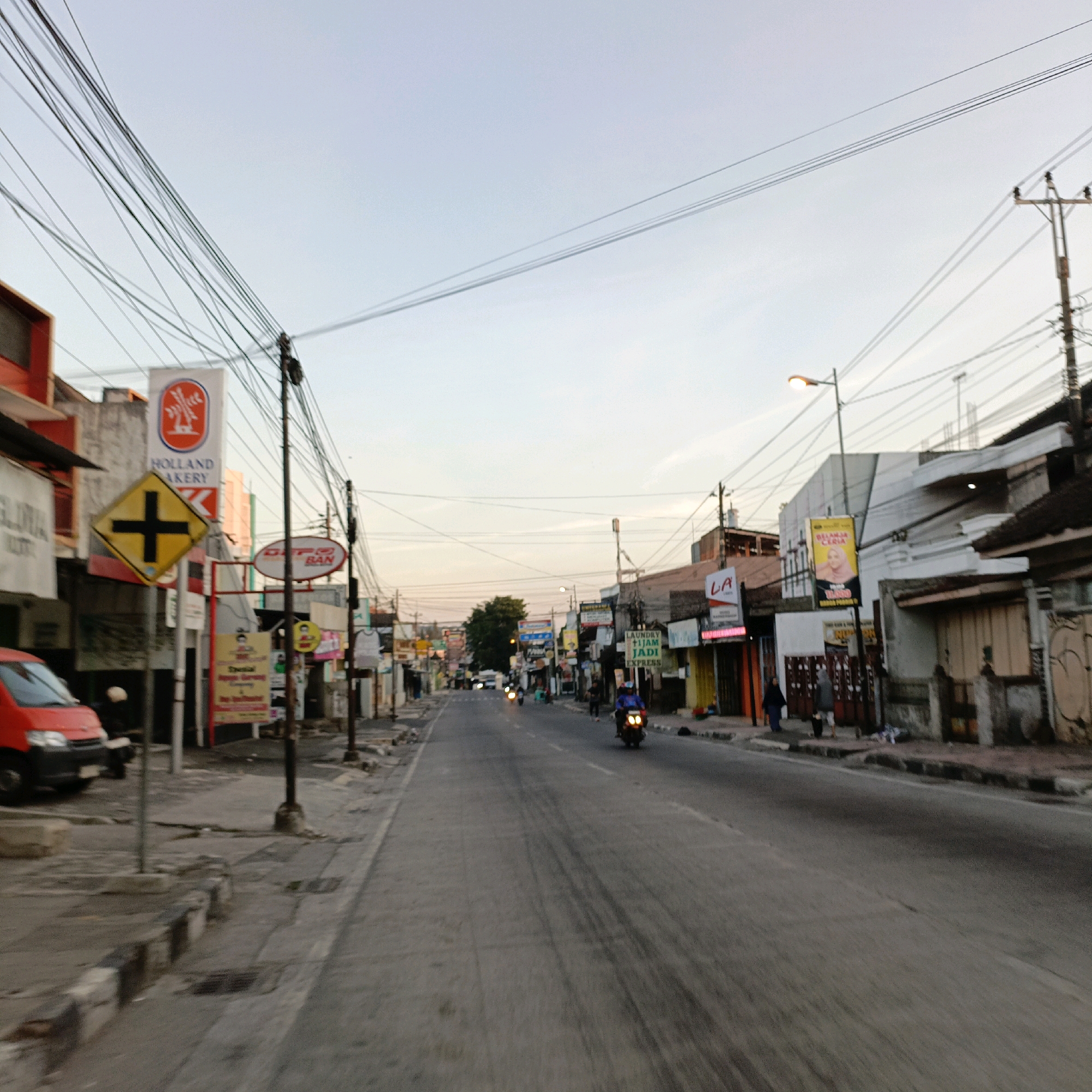 The image shows a street scene in Salatiga, Indonesia. The perspective is from a vehicle moving down the road. The street is lined with buildings on both sides, which appear to be shops or businesses. There are numerous utility poles and wires overhead, characteristic of many urban environments.
On the left side of the road, a red van is parked. A prominent sign for "HOLLAND BAKERY" is visible on a building, featuring a logo with a windmill. Below this sign is a yellow diamond-shaped traffic sign with a black cross, indicating a crossroads or intersection. Other signs in Indonesian are also present on the buildings.
The road surface shows tire marks, suggesting traffic. Several motorcycles are visible on the road, with one rider prominently in the foreground on the right, wearing a blue jacket. Other pedestrians are seen on the sidewalks. The sky is overcast with a pale blue hue, indicating either early morning or late afternoon. The overall impression is a busy, everyday street scene in an Indonesian city.