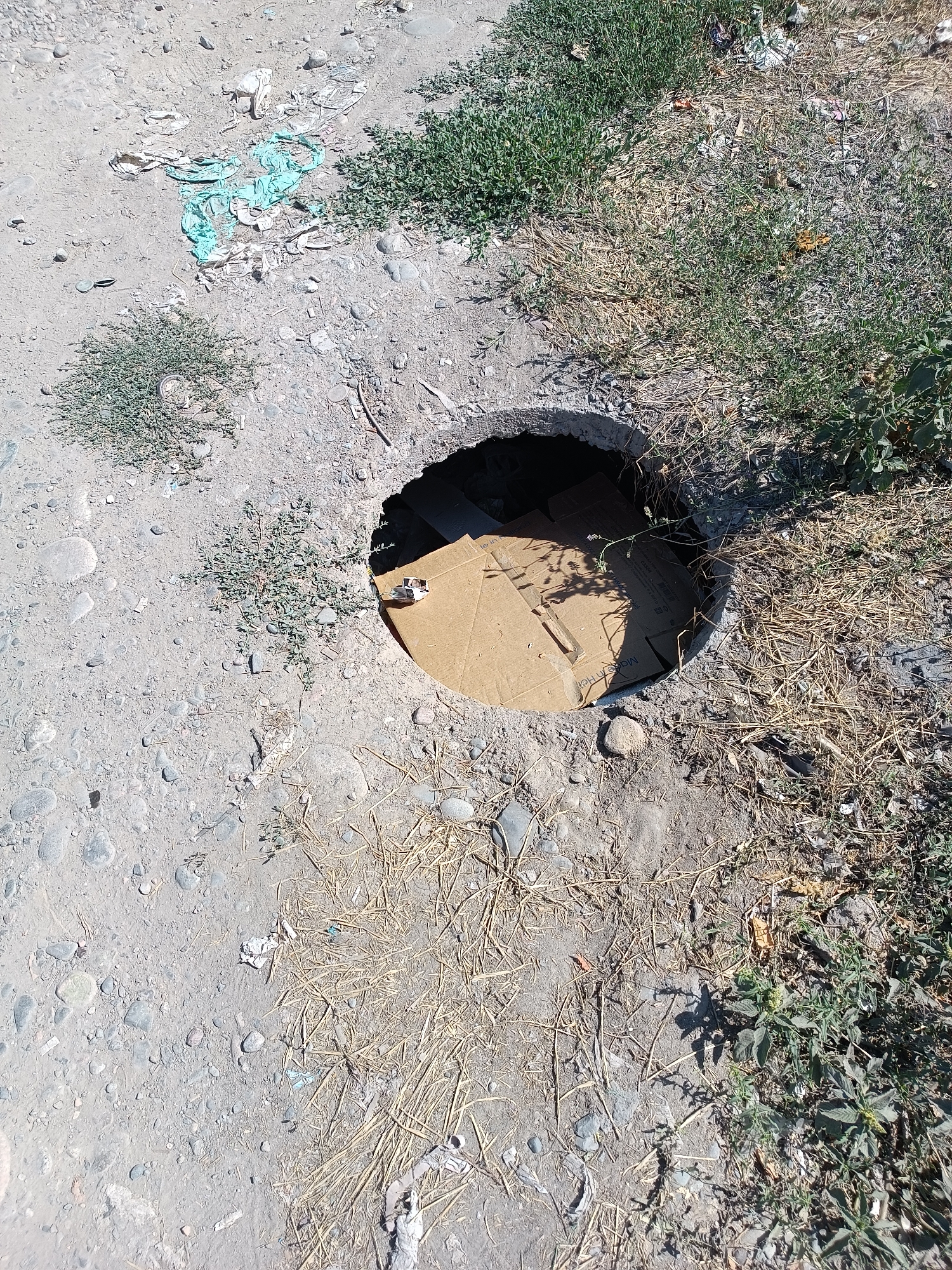 The image shows an overhead view of a broken concrete surface with a circular hole, possibly a manhole or drainage opening, that has been covered with pieces of cardboard. The surrounding ground is dry and dusty, with scattered pebbles and dried vegetation. Some debris, including plastic and paper, is visible on the ground and near the opening. The scene appears to be an outdoor urban environment, likely in a less maintained area of Bishkek City, Kyrgyzstan, due to the presence of discarded materials and the damaged infrastructure. No people are present in the image.