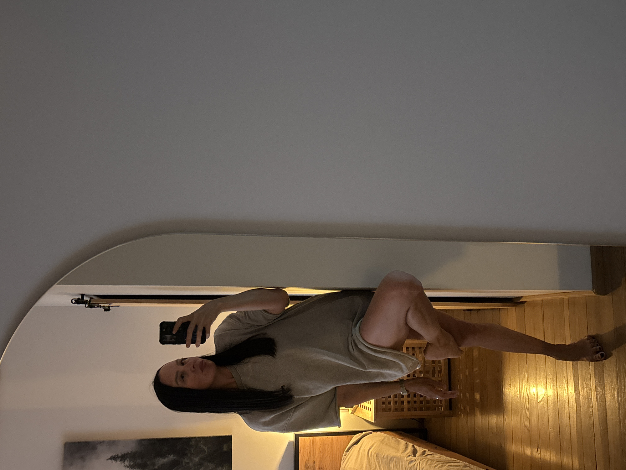 A woman is taking a selfie in a bedroom. She is wearing a light-colored oversized t-shirt and is leaning against a wall. Her right leg is bent and crossed over her left leg, and she is holding a smartphone in her right hand. A wooden bed and a nightstand are visible in the background, along with a framed picture on the wall. The floor is made of wood and reflects the warm light from a lamp. The overall scene appears to be a casual and private moment captured in a domestic setting. The location is not identifiable as Chaikovsky, Russia.
