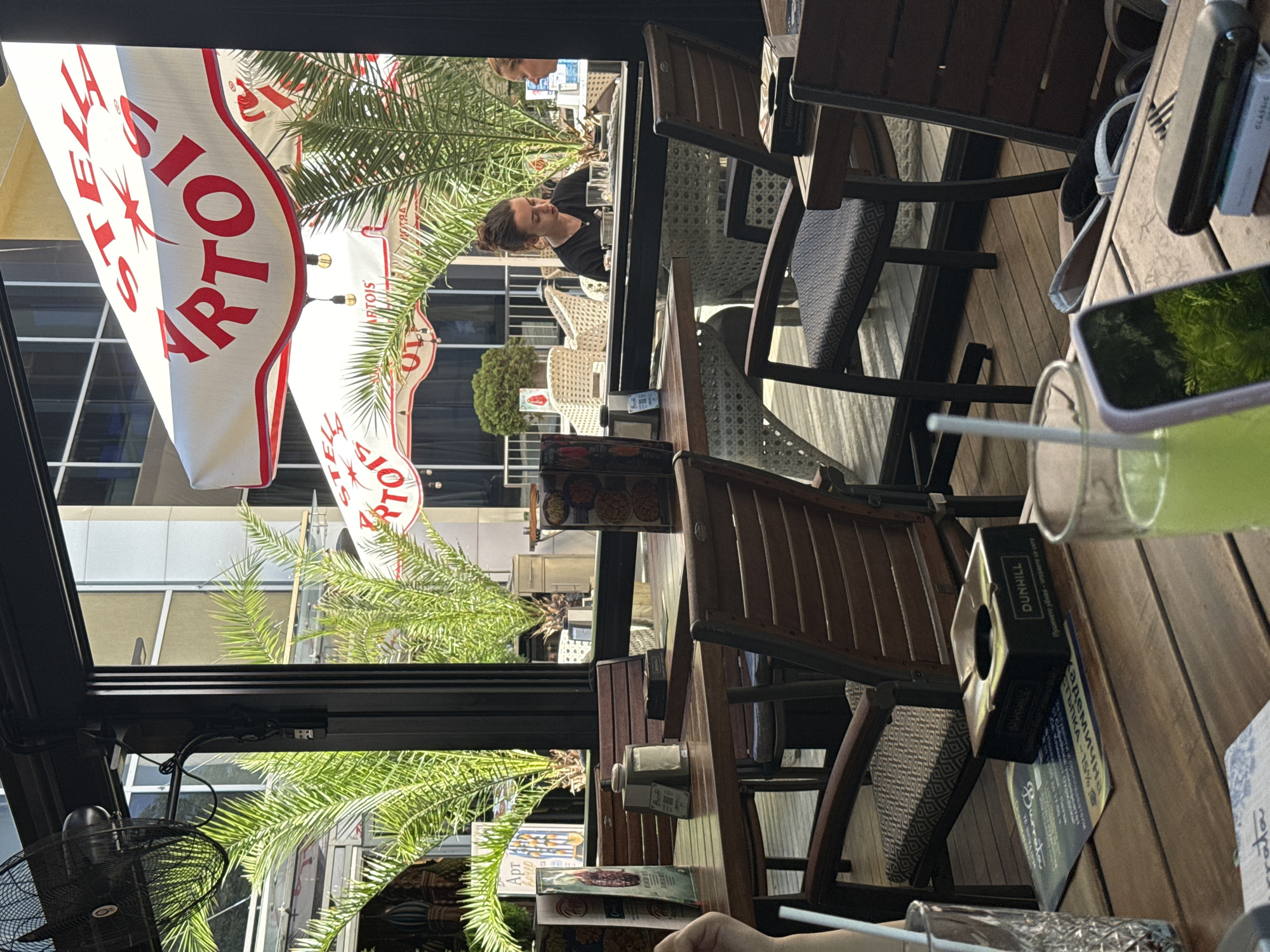 The image depicts an outdoor seating area of a cafe or restaurant. In the foreground, a wooden table is visible with several objects, including a glass with a green beverage and a straw, a smartphone with its screen reflecting greenery, a cigarette pack and lighter, and an ashtray. Behind this table, another wooden table is set with chairs.
Further back, a woman with her hair tied up is seated at a table with a white rattan chair. She is wearing a black t-shirt and appears to be looking down at something on the table. Another person is partially visible to her right. Large palm trees are present in the background, and a white umbrella with red lettering, possibly a logo, is visible overhead. The overall environment suggests a bright, sunny day. Based on the Stella Artois umbrellas and the general ambiance, this location is likely a cafe or bar in Sofia, Bulgaria, possibly a part of a chain that uses these branded umbrellas.