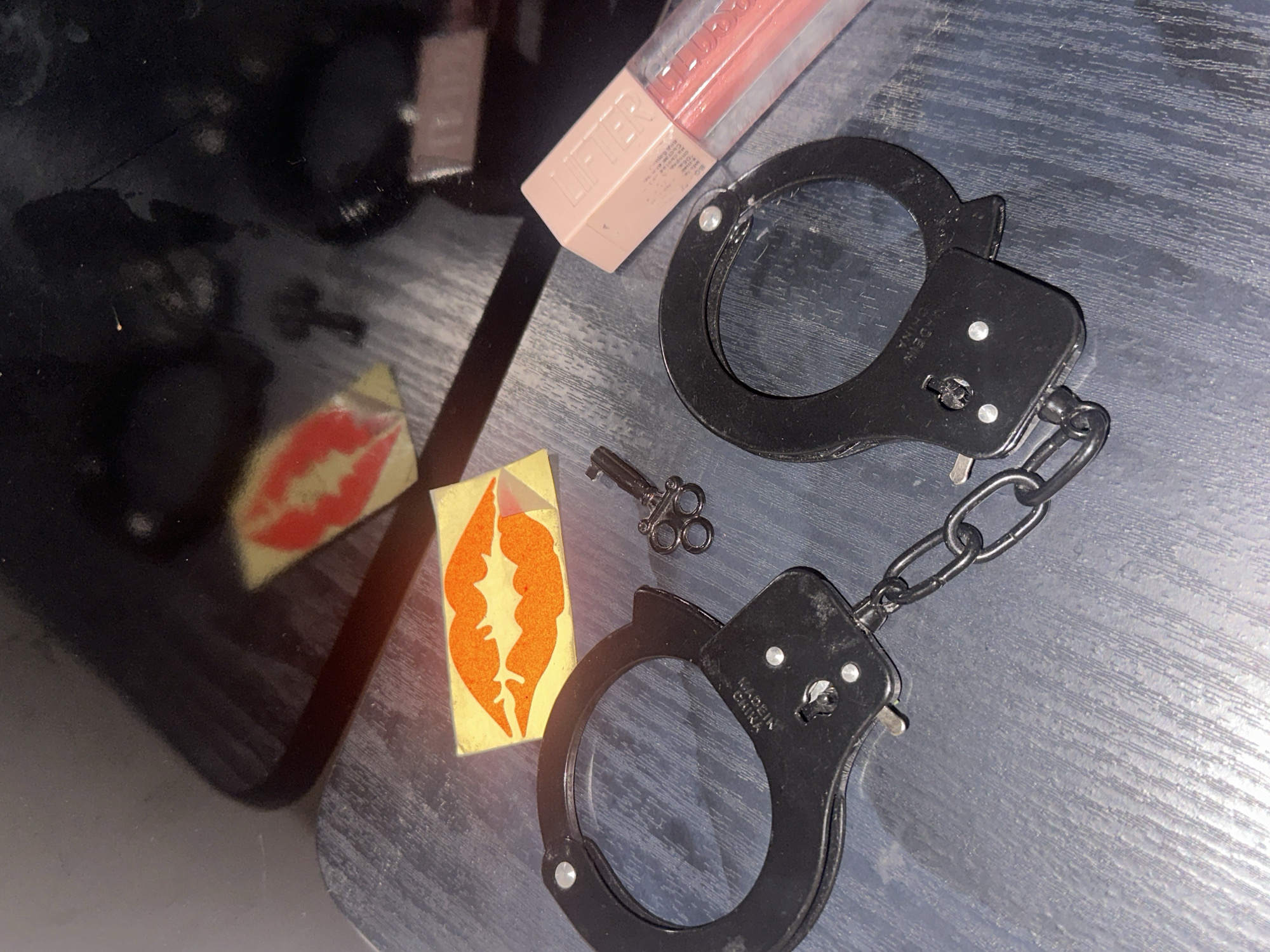 The scene, situated in the city of Sofia, Bulgaria, presents an overhead view of a dark, textured surface adjacent to a highly reflective, black tabletop. Positioned on the textured surface are a pair of black metallic handcuffs, connected by a short chain. One handcuff clearly displays "MADE IN CHINA" engraved on its face. Adjacent to the handcuffs lies a single, small, dark metallic key with an ornate, circular handle. Also on the textured surface, a rectangular paper cutout with a stylized orange lip design on a yellow background is visible.
On the reflective black tabletop, a transparent tube of peach or light pink lip gloss is present, featuring a matte pale pink cap with the embossed text "LIFTER". The brand "MAYBELLINE NEW YORK" is partially visible on the tube. Further back on this reflective surface, a second, red-colored stylized lip cutout on a yellowish background is partially obscured by reflections. The ambient lighting appears artificial, casting subtle reflections on the glossy surfaces. No individuals are present in the image.