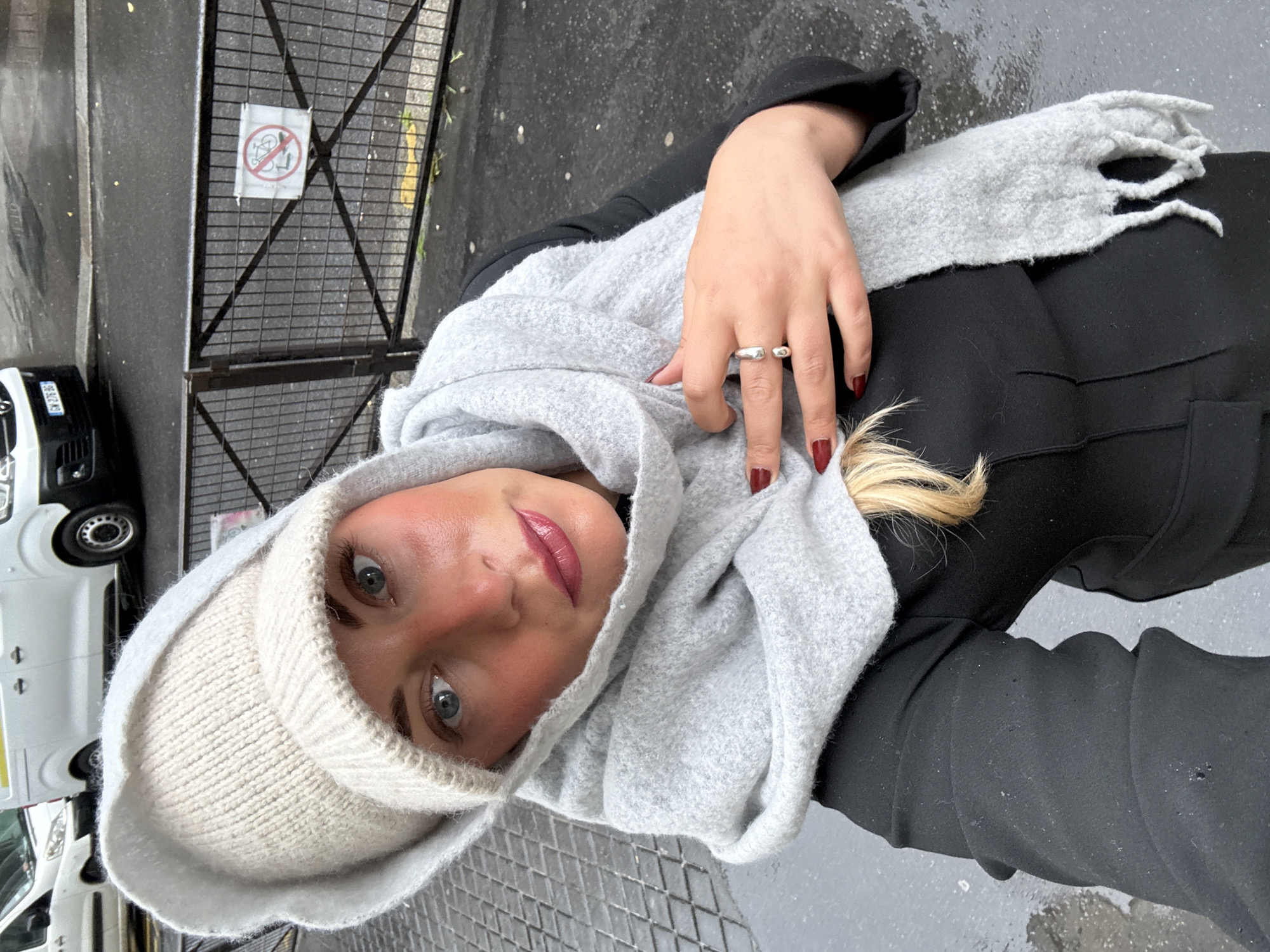 A woman in a black coat and light gray scarf and hat is standing outside in a city street on a rainy day. She is looking up at the camera, and her right hand is resting on her scarf. A silver ring is on her right index finger. Her fingernails are painted dark red. Her hair is blonde. She is standing near a fence with a sign prohibiting cycling. A white delivery van is parked nearby on the left. The location appears to be in Paris, France.