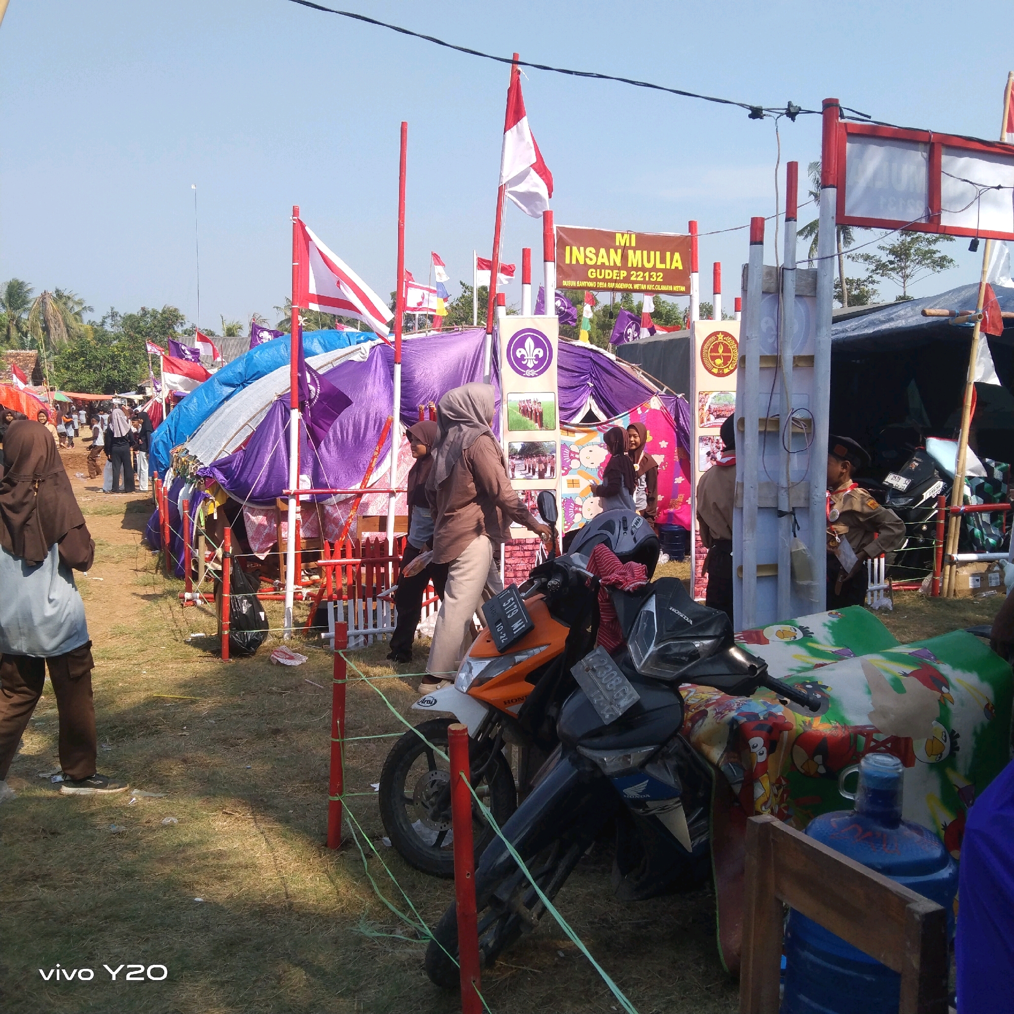 The image depicts an outdoor event, likely a scout jamboree, in Karawang, Indonesia. In the foreground, two motorcycles are parked on a grassy area. The motorcycle on the right is black, and the one on the left is orange and white. A large blue water cooler jug is visible behind the motorcycles, adorned with cartoon characters.
In the midground, several tents are set up, many covered in purple and blue tarpaulins. Red and white striped poles support the tents and fences. Flags with scout emblems and national flags are prominently displayed. A banner reads "MI INSAN MULIA GULDEP 22132 DUSUN BANTONG DESA RAJAMEMPOL WETAN KEC.CILAMAYA WETAN," indicating the name of the school, scout troop number, and location within Cilamaya Wetan subdistrict of Karawang.
Several people are present, including individuals in scout uniforms and others in casual clothing, some wearing hijabs. The sky is clear and blue, suggesting a sunny day. The overall environment appears to be a field or open space, with some trees and buildings visible in the background. The ground is covered with dry grass and scattered debris. The scene suggests an active event with participants and attendees.