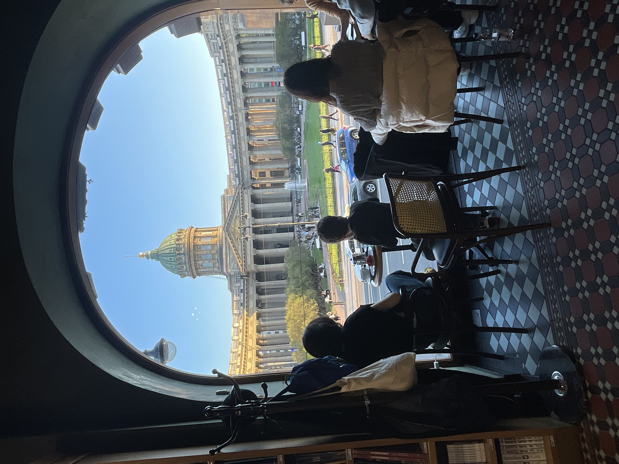 The image shows an interior scene of what appears to be a cafe or restaurant in Saint Petersburg, Russia, with a view of St. Isaac's Cathedral through a rounded window. Several patrons are seated at tables inside. The floor has a patterned tile design. Outside the window, people can be seen walking around the cathedral, and vehicles are moving on the road. The view captures a sunny day. There is a coat rack and a bookshelf in the cafe.