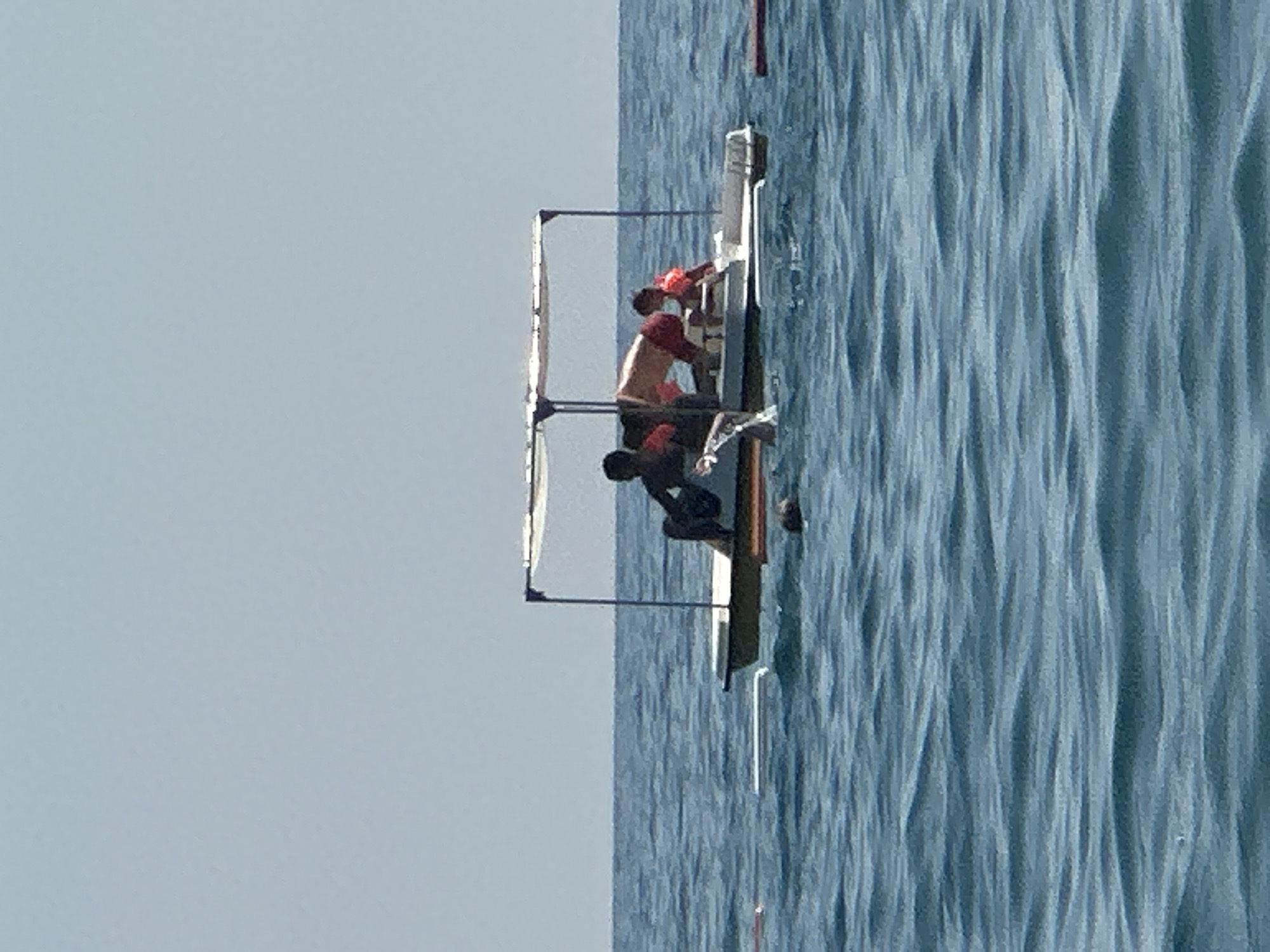 The image depicts a pedalo boat on the water with three individuals aboard. One person, appearing to be an adult, is seated on the left side of the boat. Another adult is in the center, leaning forward. A child, wearing an orange life vest, is seated on the right. The boat has a canopy providing shade. The water is calm with slight ripples, and the sky is clear and light blue. The scene appears to be on the coast of Boreti, Montenegro, a popular tourist destination known for its beaches and water activities.