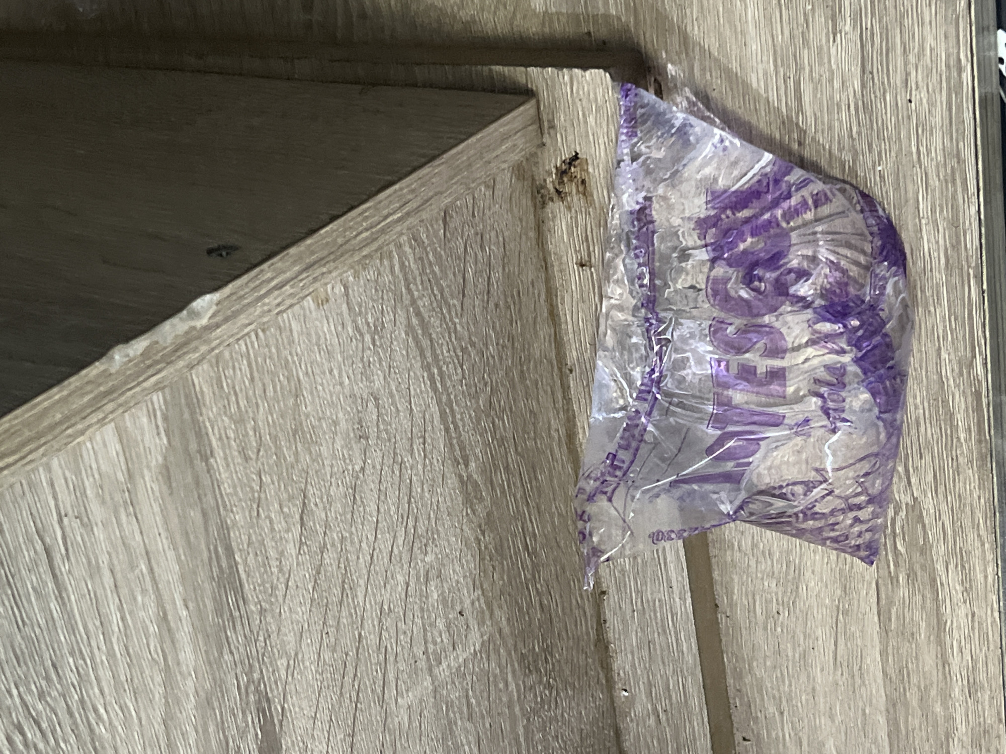 The image presents a close-up, angled view of two wooden panels or pieces of furniture joined at a corner. The wood has a light, possibly greyish-brown, hue with a prominent vertical grain texture. The upper left portion shows the underside of a shelf or panel, appearing darker.
A crumpled, translucent plastic bag with purple markings is affixed to or resting against the vertical wooden surface on the right. The bag is somewhat triangular due to its folded state. Faint purple text and design elements are visible on the bag; partial letters like "SS" and what could be part of a word like "PESS" or "SUPPLIES" are discernible. A small number or code, possibly "0230", is also visible towards the lower edge of the bag.
Minor imperfections are present on the wooden surfaces, including some rough edges and a small dark mark on the wood near the plastic bag. The scene is static, indoors, and no people or specific activities are depicted.