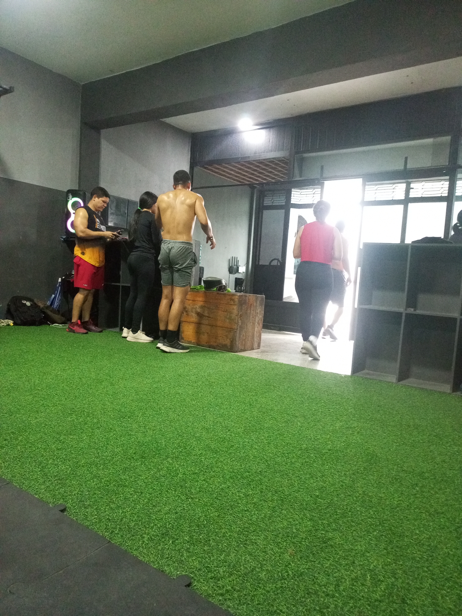 This image captures an indoor scene at a gym, likely located in Juan Griego, Venezuela, with a focus on the gym floor covered in artificial turf. Four individuals are present. On the left, a man wearing a yellow and red tank top and red shorts is looking at his phone. Next to him, a woman in black athletic wear stands beside him. To their right, a shirtless man in grey shorts is facing away from the camera, appearing to be mid-conversation or interaction. Further back, a woman in a pink top and black leggings walks away from the camera, entering a brightly lit doorway. The environment features gym equipment and storage shelves in the background, along with a wooden crate. The lighting suggests an indoor setting, possibly with natural light from the doorway.