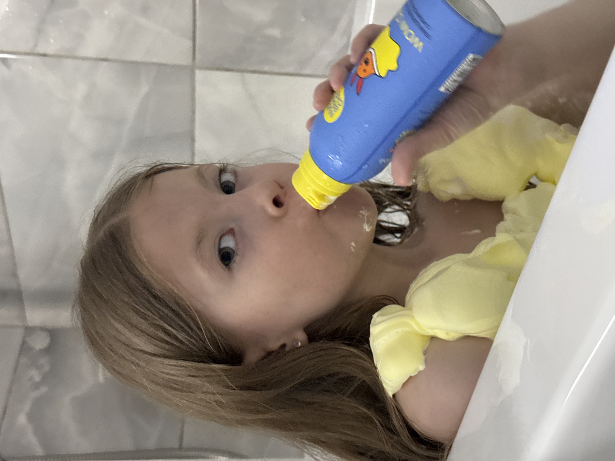 A young girl is in a bathtub, possibly a bathtub in a bathroom with white tiles. She has brown hair and is wearing a yellow garment. She is holding a blue bottle with a yellow cap up to her nose/mouth, possibly ingesting or applying something from the bottle. The bottle has cartoonish drawings on it. The exact location in Izhevsk, Russia cannot be determined from the image.