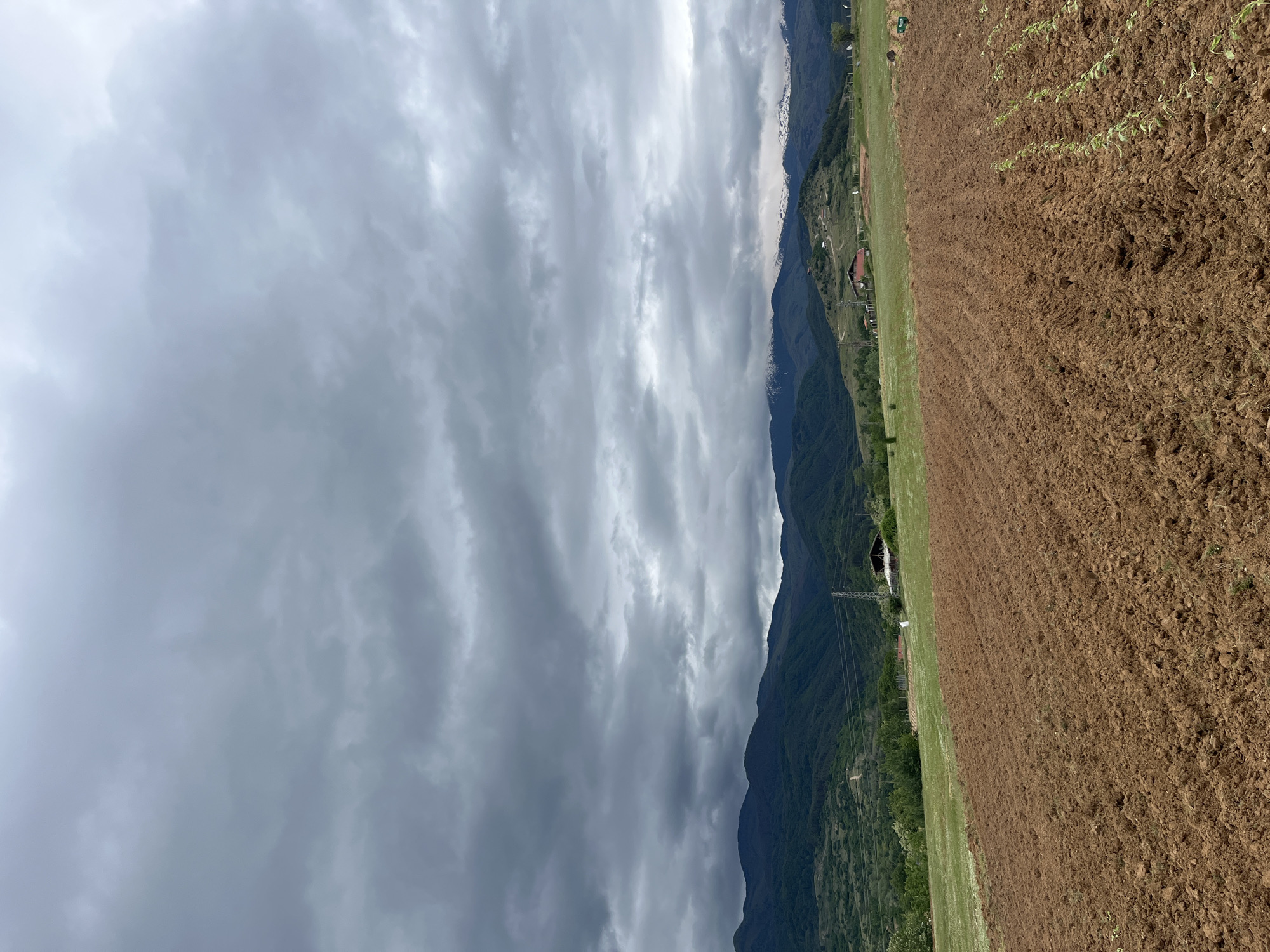 The image depicts a landscape in Kornitsa, Bulgaria. On the right, there is a field of tilled brown soil with some young green plants emerging. Adjacent to the field is a strip of green grass and some sparse vegetation. A mountain range covered with trees and some structures runs along the center of the image. Transmission towers are also visible near the mountains. The sky is filled with thick, gray clouds, suggesting an overcast or stormy weather.