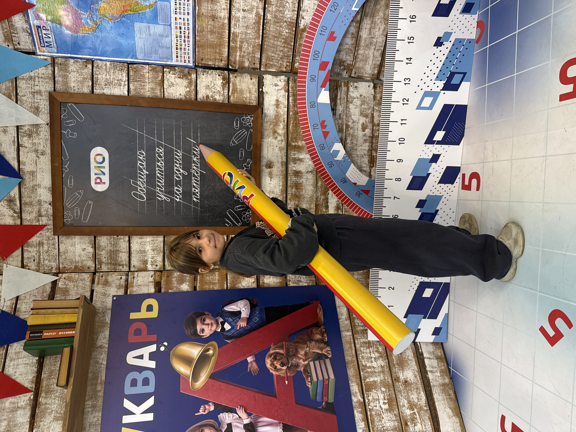A young girl, approximately 7-9 years old, stands in a brightly decorated room that appears to be a back-to-school or children's educational display. She is holding a large, oversized yellow pencil with red ends, pointing it towards a blackboard. The blackboard features chalk drawings and text in cursive, including the word "РИО" within a logo. The girl is wearing a dark gray sweatshirt and dark pants.
The room's walls are made of distressed wooden planks. To the girl's left, a large blue banner with the Cyrillic letters "УКВАРЬ" and an illustration of children and a dog is visible. To the girl's right, a world map and a large protractor-like ruler with numerical markings are displayed. There are also triangular paper flags in red, blue, and white strung across the top of the scene. The floor has a grid pattern with large red numbers.
The scene suggests a playful and educational environment, possibly a photo zone for children. There are no indications of the city of Ignatovo, Russia, within the visual elements of the image.