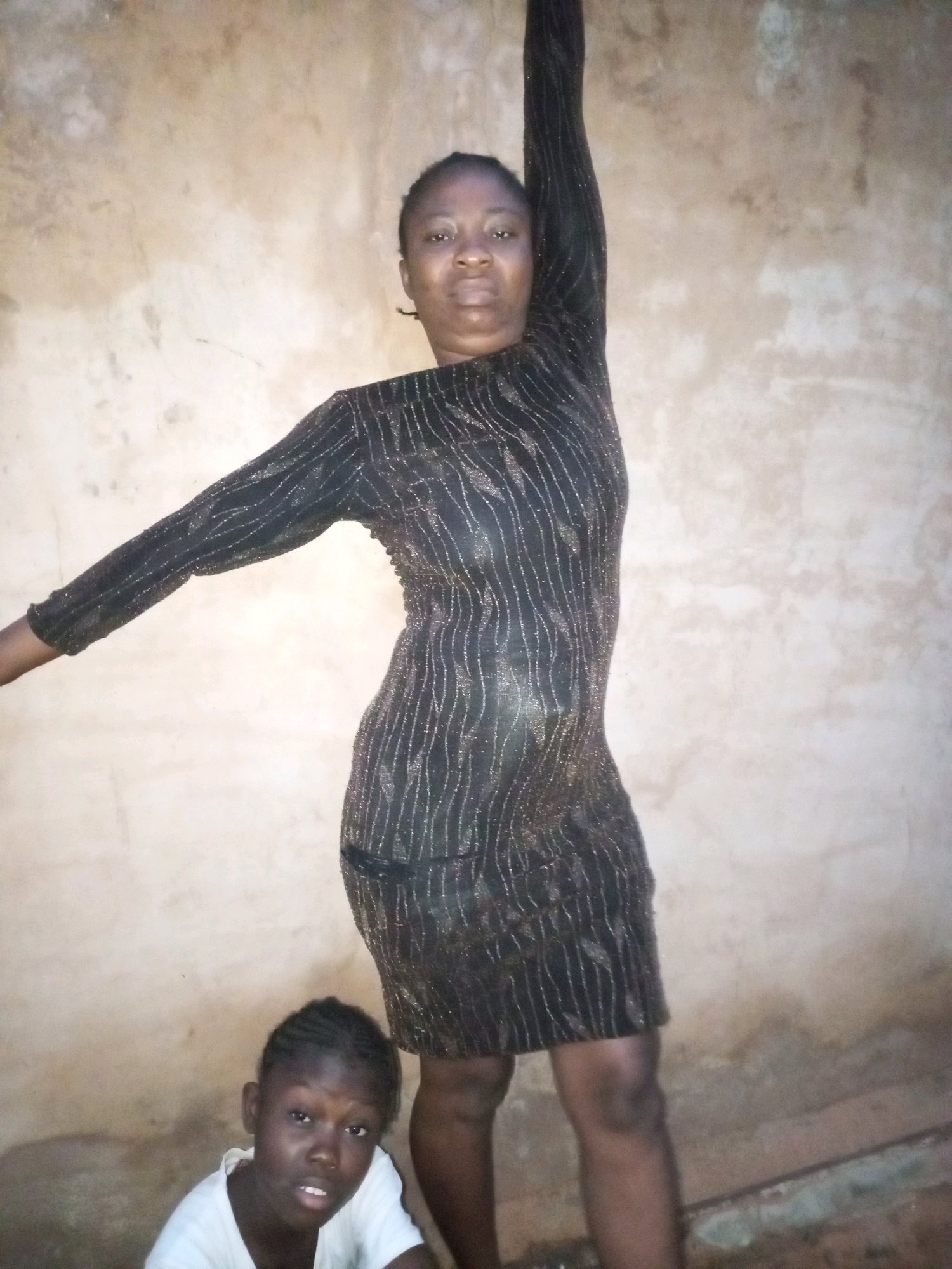 In a room with a textured, pale beige wall, a woman stands to the right. She is wearing a long-sleeved black dress with vertical, shimmery lines. Her right arm is raised high above her head, and her left arm is bent at the elbow, extending forward. To the left and in front of the woman, a young girl sits on the floor. She is wearing a white t-shirt. The room appears to be a simple indoor space, possibly in Tempure, Nigeria.