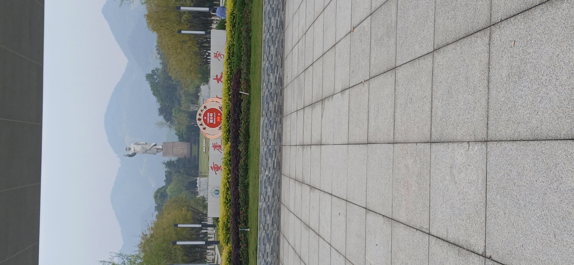 The photograph depicts a paved area adjacent to a green strip and signage, with a statue visible in the background. The location is Chongqing High-tech Zone, China. A concrete or stone tiled pavement makes up the foreground. Adjacent to the pavement is a strip of greenery with bushes. Signage with Chinese characters is also visible in the greenery. In the background is a statue set against a backdrop of mountains. The sky is overcast. No people are clearly visible.