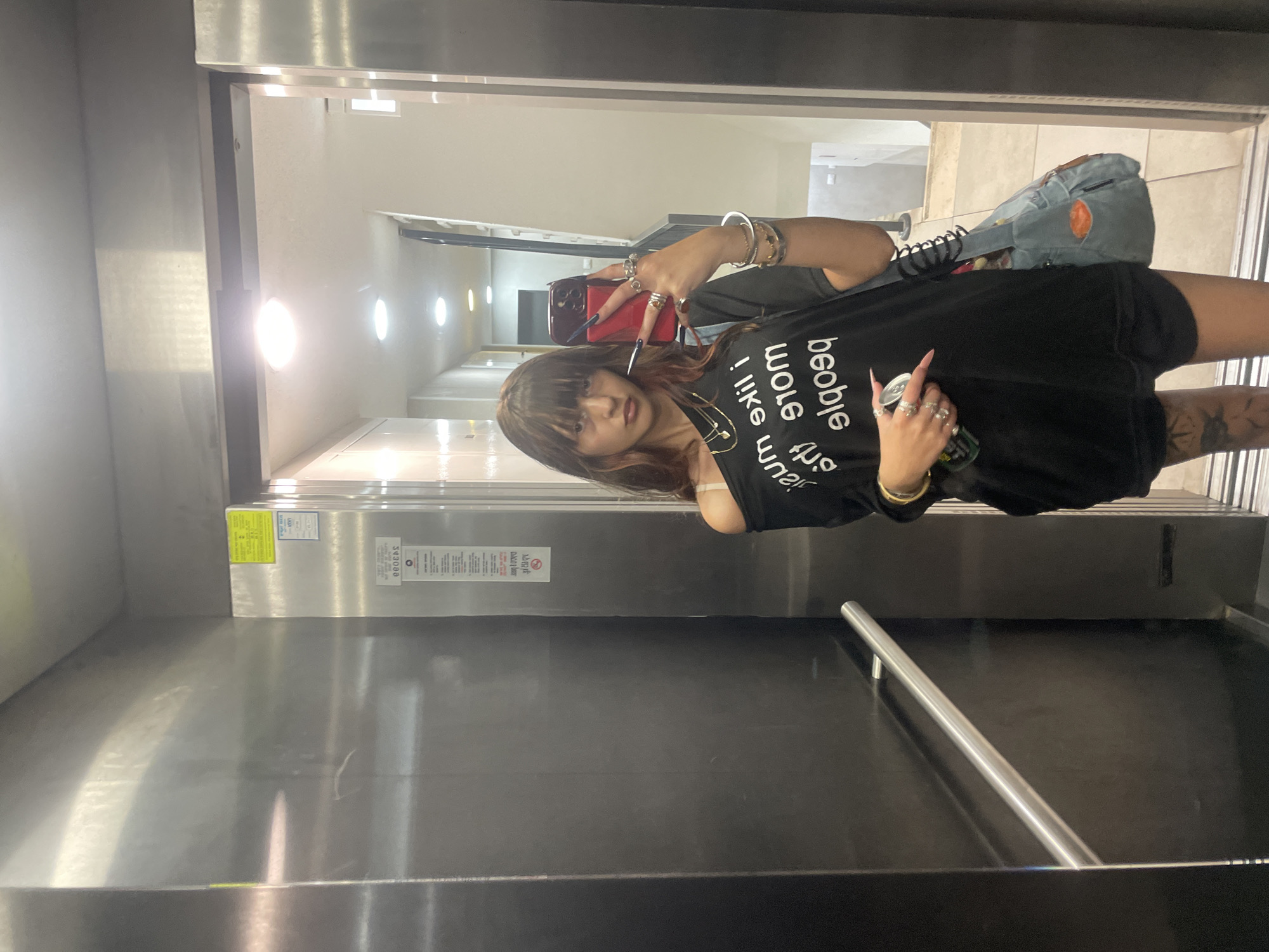 A young woman with bangs and dyed hair takes a selfie in an elevator. She is wearing an off-the-shoulder black t-shirt with white text that reads "I like music more than people," layered necklaces, a bracelet, and several rings on her fingers. She is holding a red cell phone in her right hand and a can of soda in her left. She is also wearing black shorts and has tattoos on her legs. The elevator walls are made of brushed metal, and there is a handrail on the left side. A sign with text in Hebrew is visible on the right wall of the elevator. The elevator appears to be located in an apartment building in the city of Raanana, Israel, as indicated by the text on the sign.