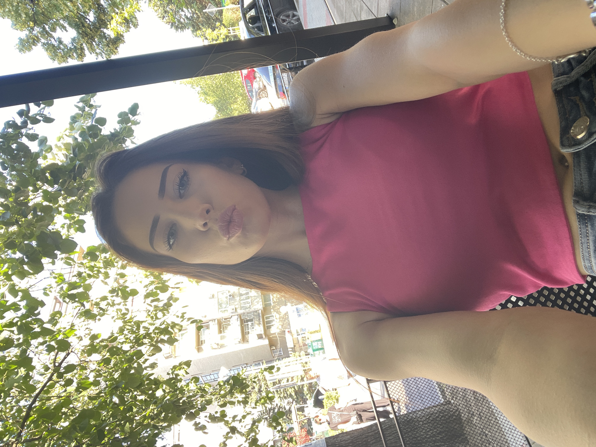 An outdoor, medium-shot selfie features a young woman with long brown hair and a pink tank top, pouting her lips. She is sitting outside, with trees and buildings visible in the background. The scene appears to be in a city.