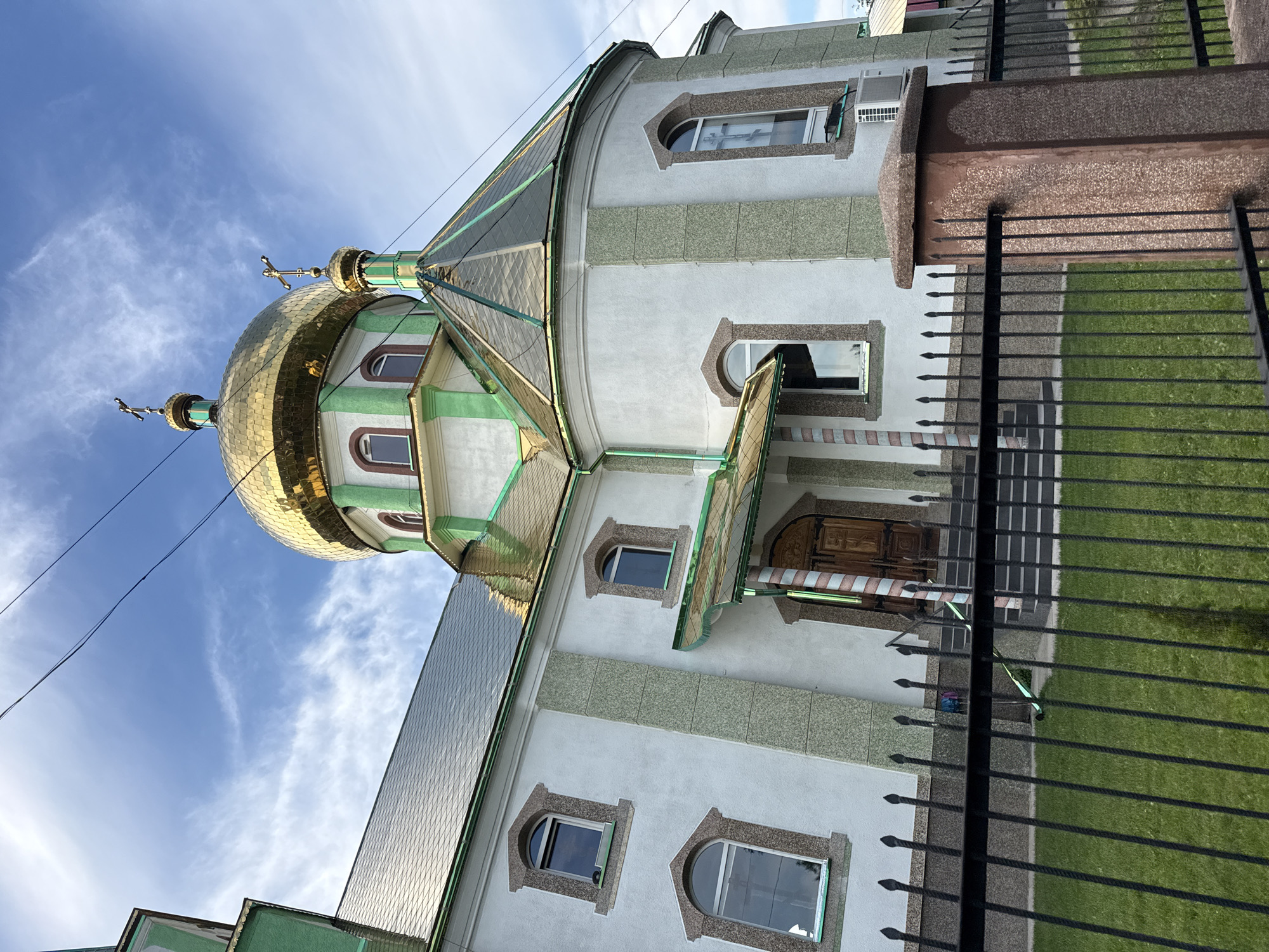 The image shows the exterior of an Orthodox church with a golden dome, located in the city of Ilnytsia, Ukraine. The church is white with green accents and has arched windows. A black metal fence is in the foreground, with a well-manicured green lawn behind it. The sky is blue with scattered clouds. There are no people visible in the image.