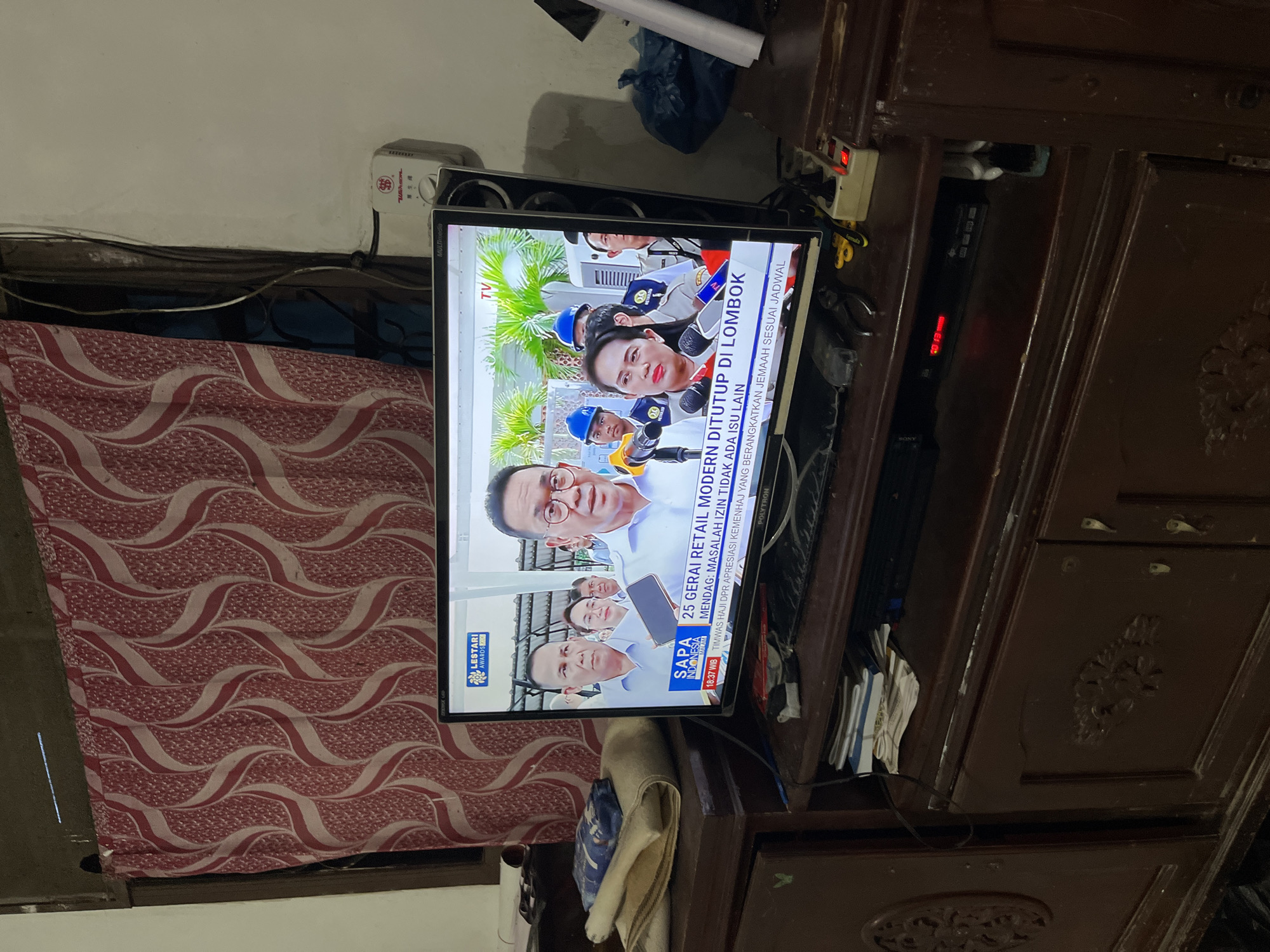 A television displays an Indonesian news broadcast featuring images of people in suits and political figures, with text captions in Indonesian. The TV is placed on top of a dark, ornate wooden cabinet. A patterned curtain is visible to the left of the TV. A white extension cord and other miscellaneous items are in the background. The broadcast mentions the closure of modern retail outlets in Lombok. The TV screen shows the time as 18:37 WIB and the news program title as "SAPA INDONESIA." The location is possibly Keudematangkuli, Indonesia.