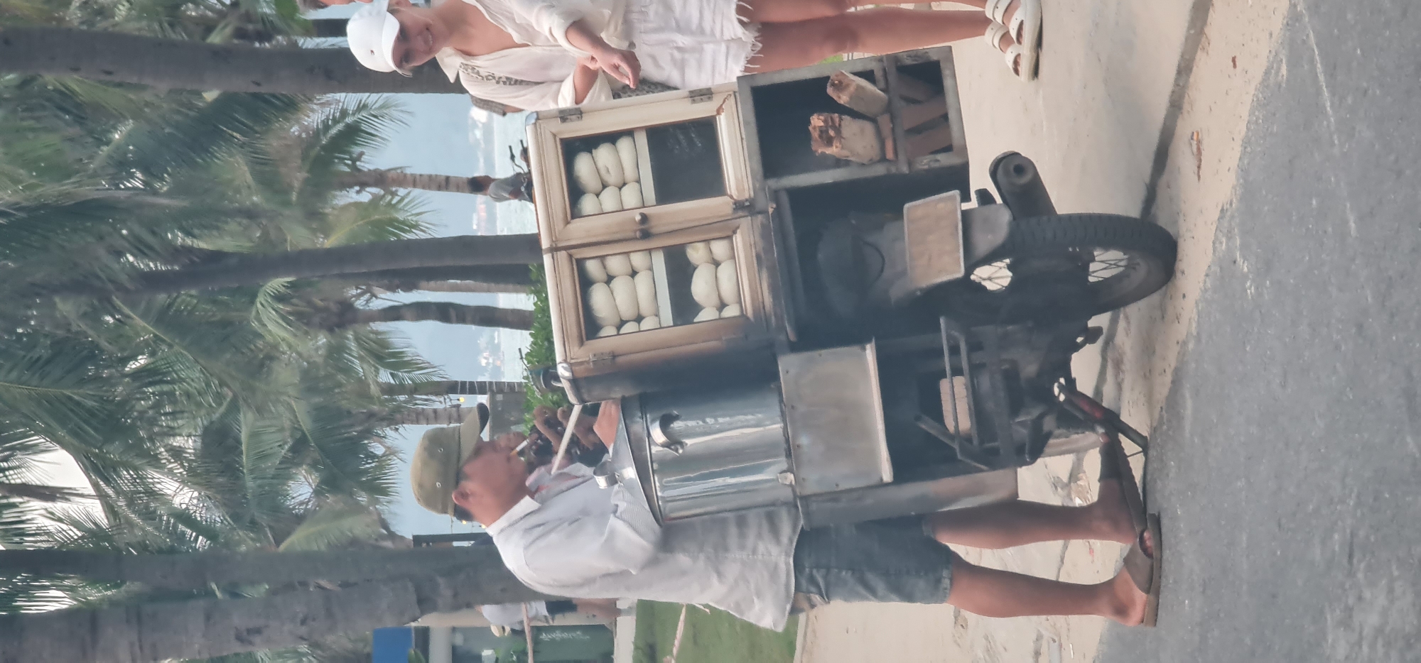 This photograph captures a common street food vending scene, likely in Đà Nẵng, Vietnam, during the late afternoon or early evening. The main subject is a male street vendor, dressed in a light-colored long-sleeve shirt, dark shorts, and a cap, standing barefoot in sandals. He is seen holding a lit cigarette, appearing to be attending to his mobile food cart.
The food cart is a custom-built setup attached to a motorcycle. It features a prominent wooden display cabinet with glass panels, showcasing rows of white, oval-shaped food items, most likely steamed buns (bánh bao or similar). Above this display, an open compartment holds several pieces of firewood or charcoal, suggesting it's used to fuel a cooking or heating apparatus within the cart, possibly a steamer or oven, whose metallic parts are also visible. The motorcycle itself appears to be a small, older model.
Standing opposite the vendor is a woman, partially visible, dressed in a white outfit with a white hat and white sandals. She appears to be a customer, looking down at the food on display.
The background is rich with tropical foliage, primarily tall palm trees with their distinctive fronds. Beyond the trees, a tranquil body of water, likely the sea, stretches towards the horizon, meeting distant hills or mountains under a soft, light blue sky. This coastal and green environment aligns perfectly with the specified location of Đà Nẵng. The scene depicts an everyday interaction of commerce and food culture, with no strong emotions discernible. There is no visible text in the image.