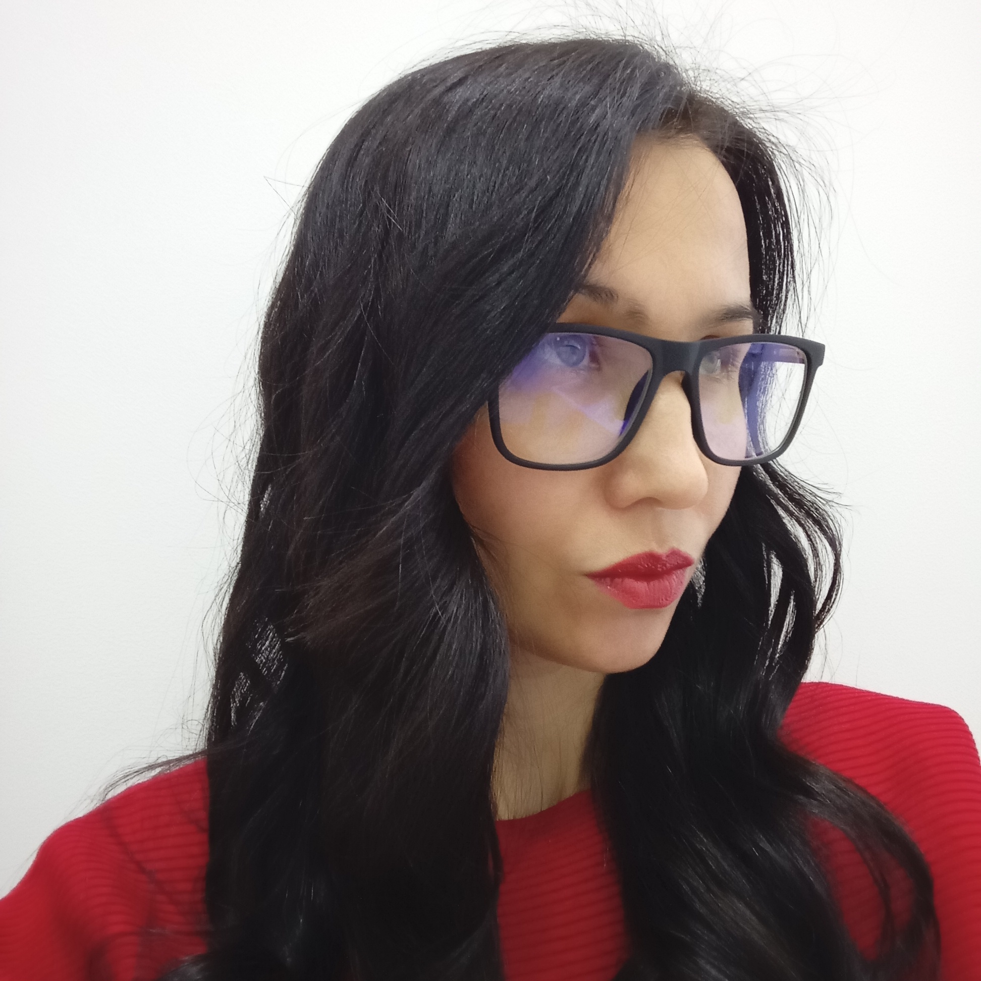 A woman with long, dark hair and red lipstick is wearing black-framed glasses and a red ribbed shirt. She is facing slightly to the right and looking away from the camera. The background is a plain white wall. The location cannot be determined from this image.