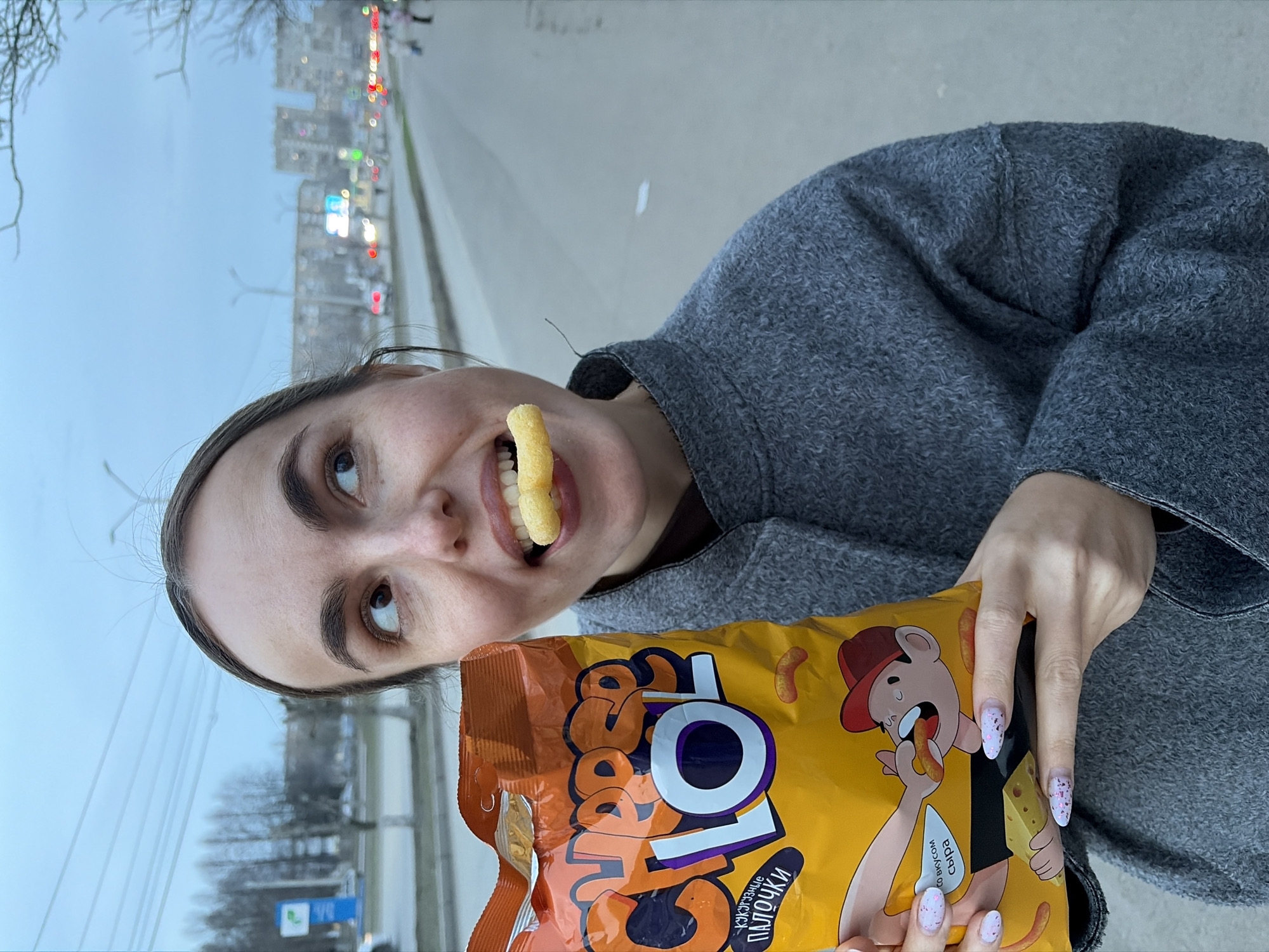 A young woman stands outdoors, holding a bag of "CRAB'S LOL" corn sticks in front of her. She has one corn stick held in her mouth. She is wearing a gray coat. The background shows an urban scene with buildings, streetlights, and a road, identifying the location as the city of Izhevsk, Russia.