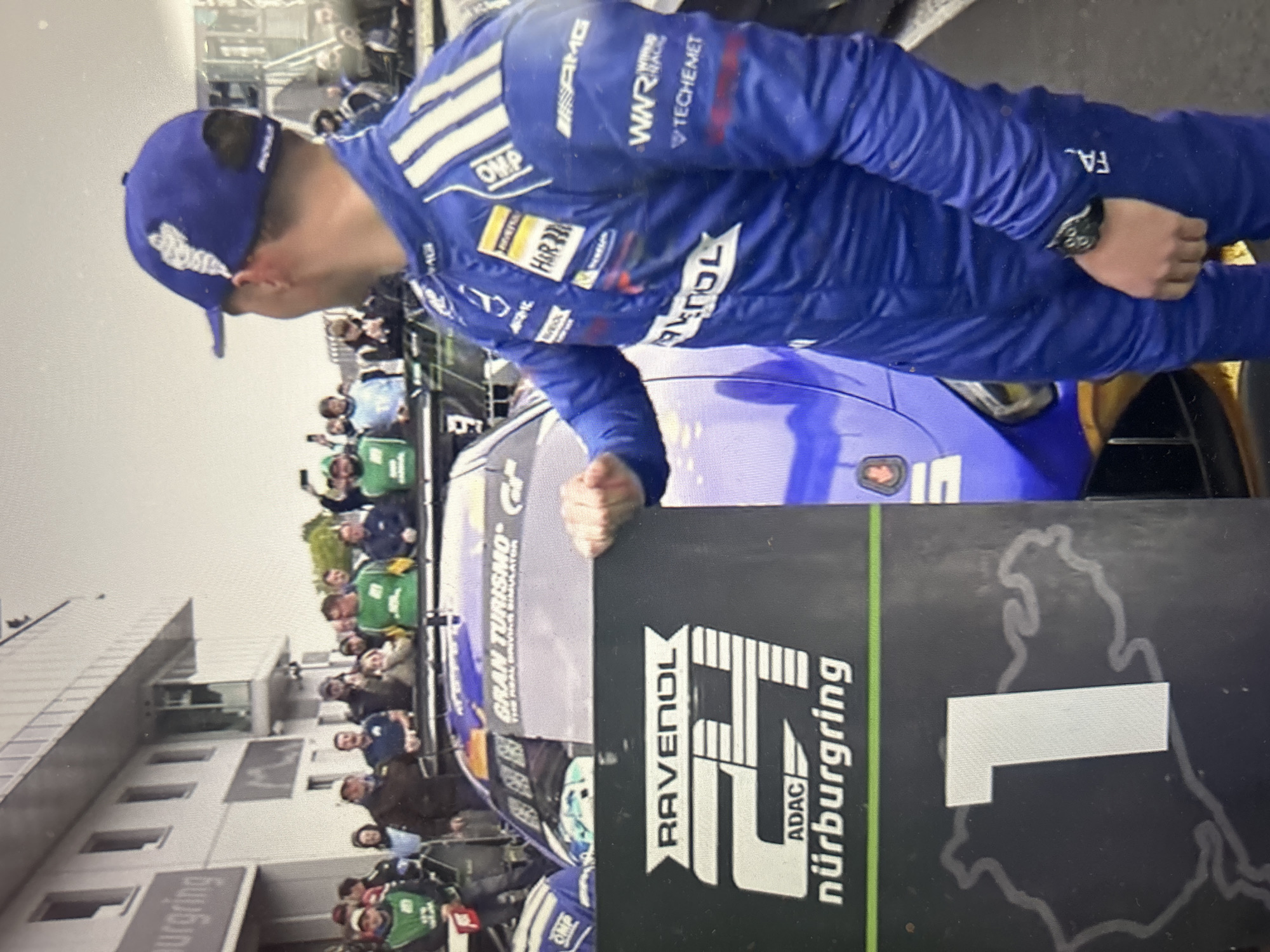 A racing driver in a blue uniform and matching cap is posing next to a sign that reads "RAVENOL, ADAC, nürburgring" and includes the number "1." The driver has several sponsor logos on their uniform, including "AMG," "OMP," "H&R," "WWR," and "TECHEMET." There is a race car partially visible behind the sign, and a crowd of people is visible in the background. The building in the background bears the name "nürburgring." The letters "FA" are on the sleeve of the blue uniform. The sign also has "GRAN TURISMO" on it. The scene is not in Sofia, Bulgaria, but at the Nürburgring racing complex.