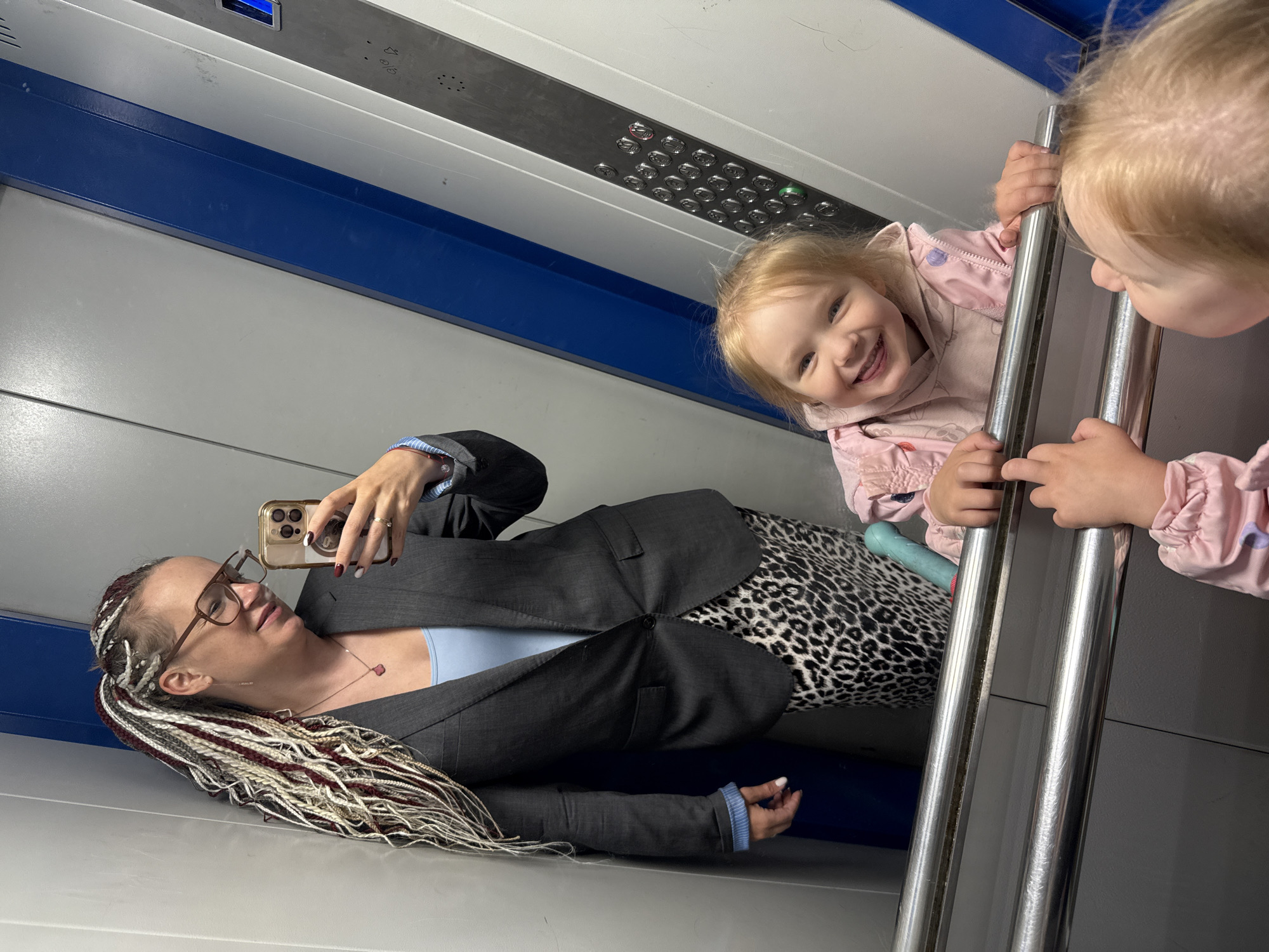 In an elevator in Moscow, Russia, a woman with braided hair in white, grey, and maroon, wearing glasses, a grey blazer, a light blue top, and a leopard print skirt, takes a selfie. In the reflection of the elevator doors, a young girl with blonde hair, wearing a pink hooded jacket, smiles and sticks out her tongue, leaning on the elevator railing. Another young child's head is visible in the foreground reflection, also looking towards the girl. The elevator interior features grey and blue paneling and a control panel with buttons.