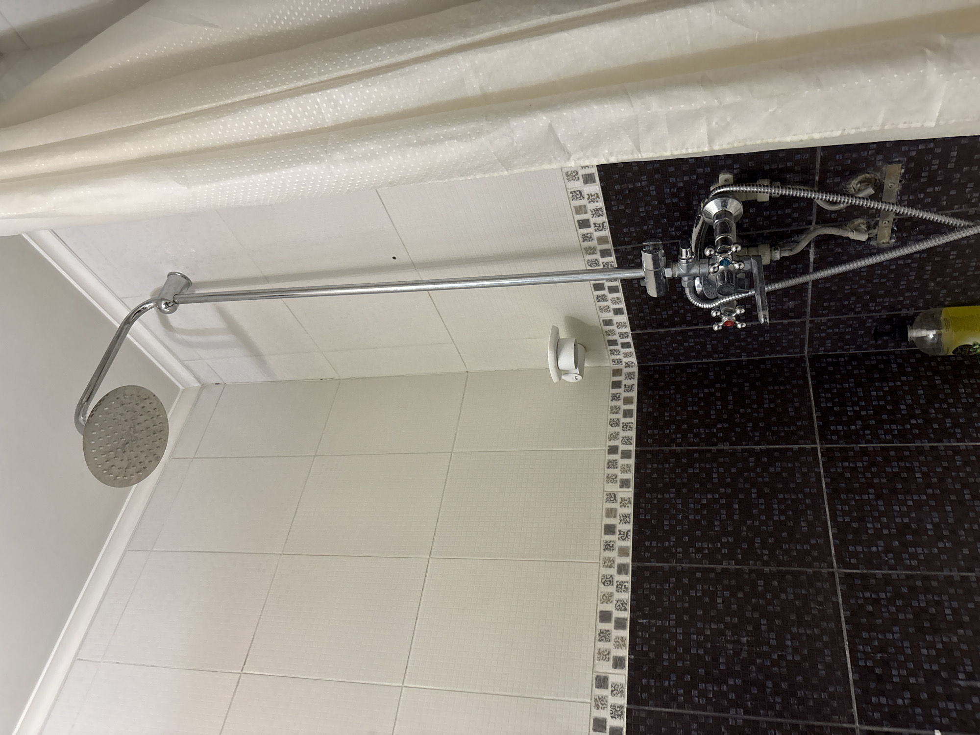 A shower stall is depicted, featuring a chrome shower head mounted on a long chrome pipe extending from a shower mixer. The stall is tiled with light beige tiles on the upper section and dark brown mosaic tiles on the lower section, separated by a decorative border of smaller patterned tiles. A white shower curtain hangs to the right. A white soap dish is attached to the wall above the shower mixer. The shower mixer itself is chrome and has red and blue indicators on its knobs, suggesting hot and cold water controls. Hoses are connected to the mixer. Below the mixer, a bottle of yellow liquid, likely shampoo or body wash, is visible on the floor. The scene is likely an indoor bathroom. There is no discernible information in the image that would indicate its specific location in Shymkent, Kazakhstan.