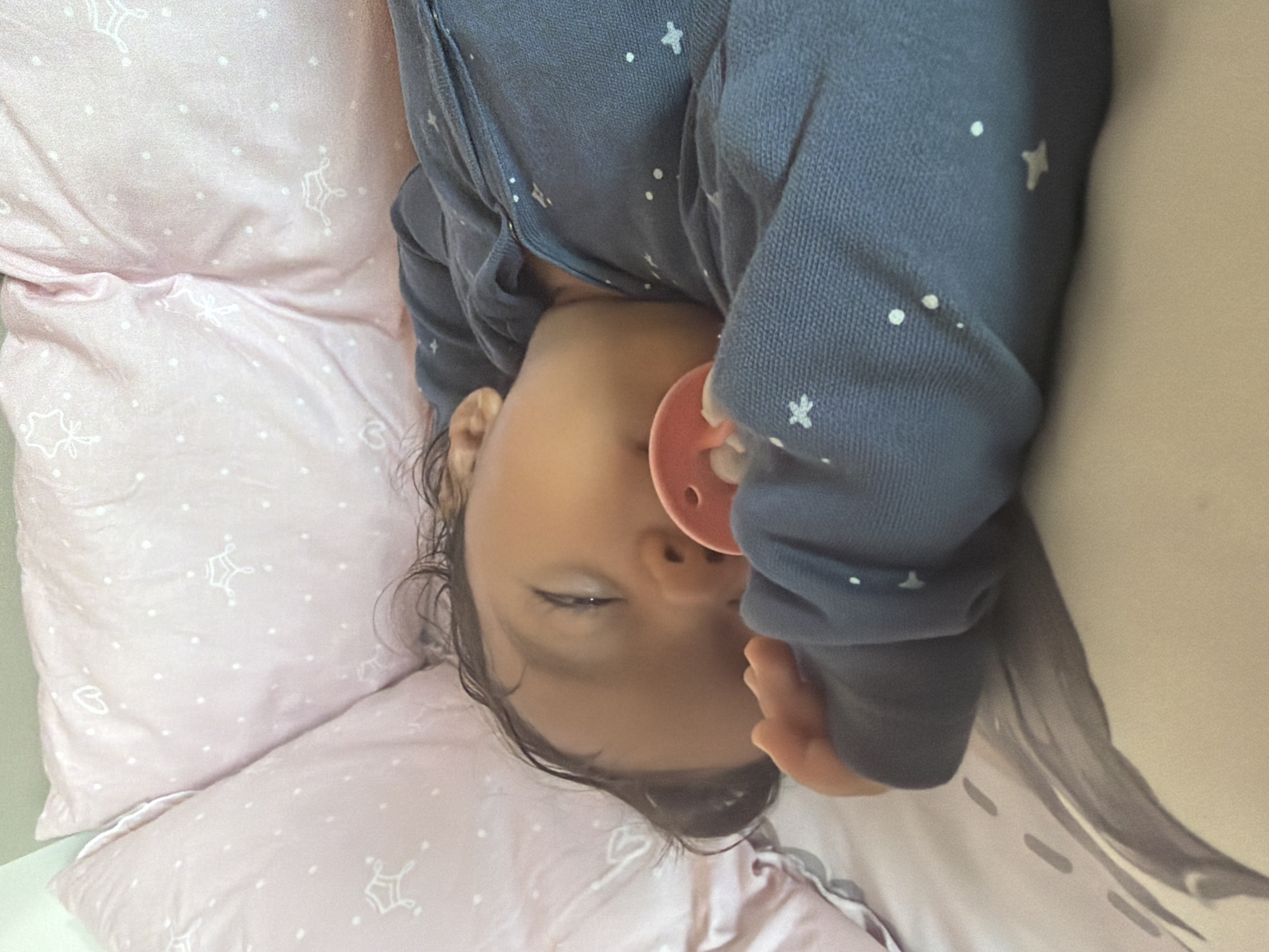 A young child is sleeping on their side in a crib. The child is wearing a dark blue outfit with white stars. A pink pacifier is in the child's mouth. The crib is covered with pink pillows with white star patterns and a blanket with abstract gray designs. The image was taken in Razhevo Konare, Bulgaria.