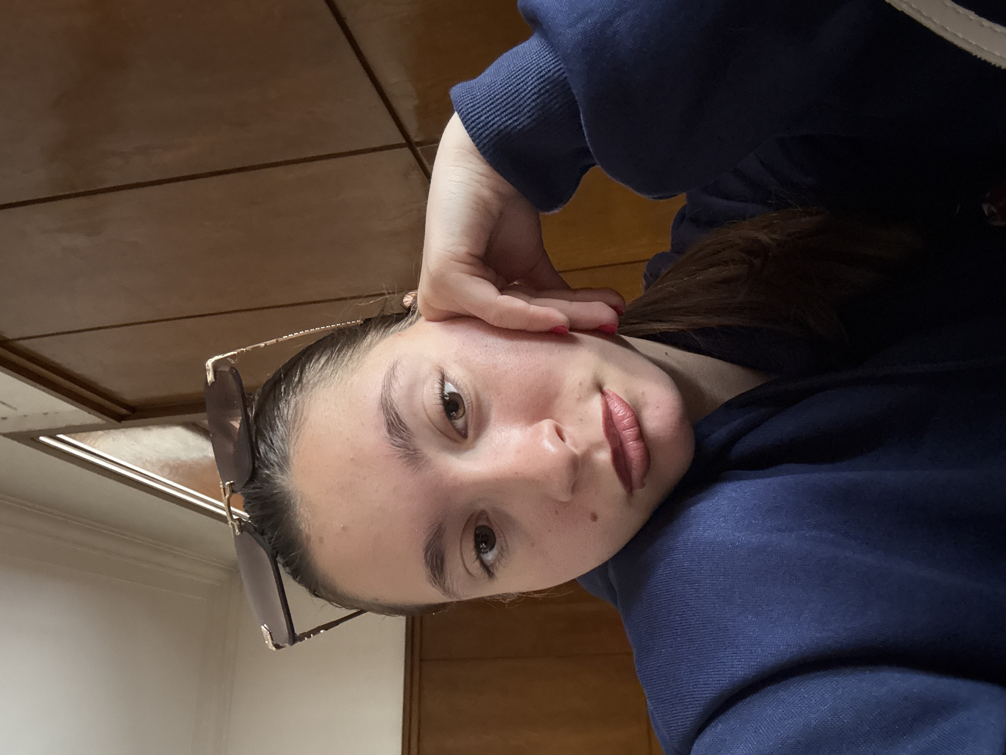 A young woman is pictured in a close-up, slightly canted selfie. She has brown hair pulled back, brown eyes, and fair skin. She is wearing a navy blue hooded sweatshirt and has sunglasses perched on her head. Her left hand is near her face. The background is a wooden surface and a white wall. The location and city are unknown.