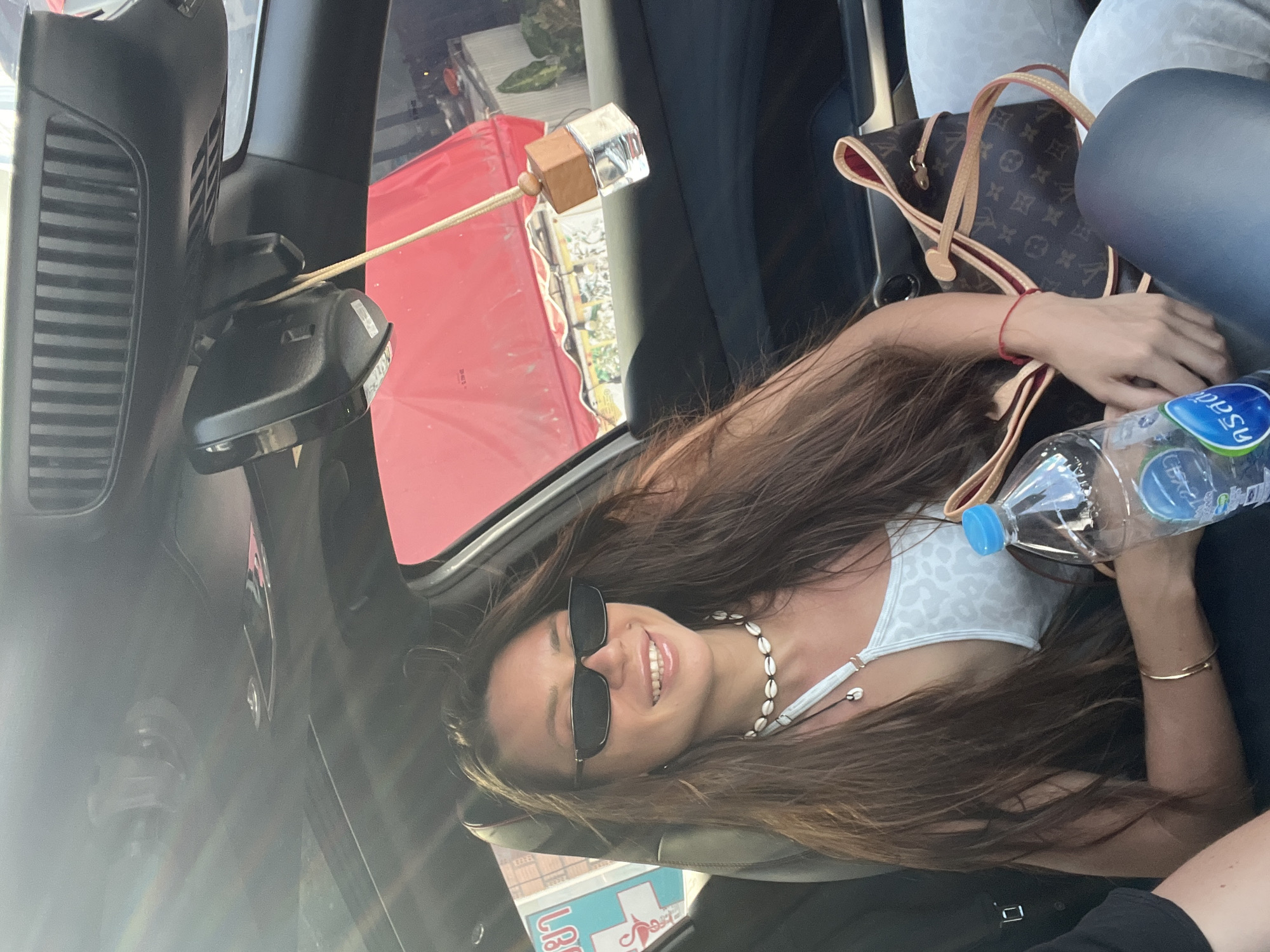 The image shows a woman seated in the passenger seat of a car, facing the camera and smiling. She is wearing sunglasses, a patterned top, and a shell necklace. Her long hair flows around her shoulders. She is holding a water bottle. A Louis Vuitton handbag sits beside her.
The car's interior is visible, including the dashboard and rearview mirror. Hanging from the rearview mirror is a small, decorative air freshener.
Outside the car window, a red awning is visible, along with signs in Thai script. The image suggests the location is บ้านบางรักษ์, Thailand, as indicated on one of the signs visible in the background.