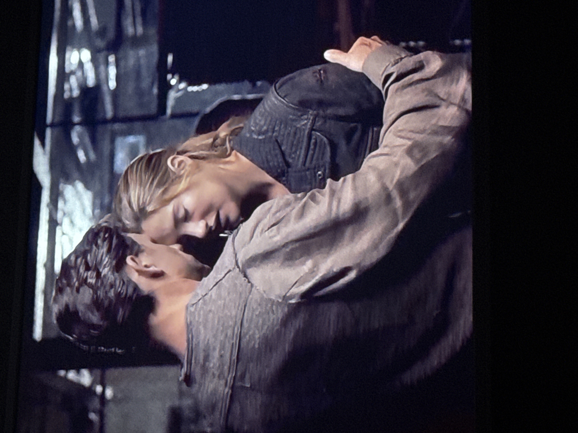 The image depicts a man and a woman embracing and possibly kissing. They are both wearing dark-colored clothing. The man has short, dark hair and is facing away from the camera. The woman has blonde hair and is facing toward the camera. The background is indistinct and appears to show a window with some objects outside. There is no indication of the city of Mytishchi, Russia in the image.