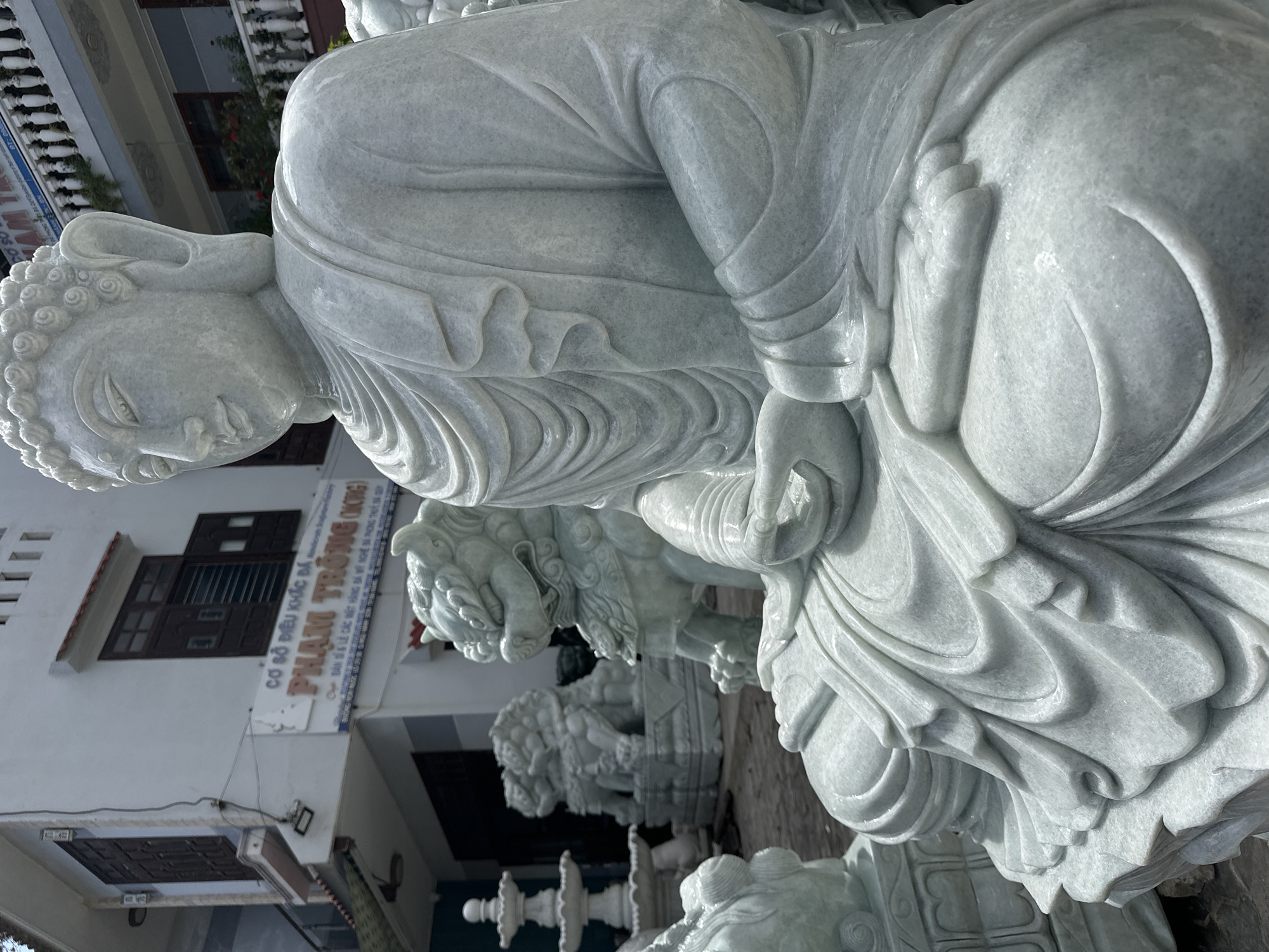 The image shows a large, intricately carved white marble statue of Buddha in a seated position. The Buddha statue is the primary subject, with its detailed robes and serene facial expression. In the background, there are other stone sculptures, including a fountain and several lion statues. A building with a sign in Vietnamese is visible, which reads "CƠ SỞ ĐIÊU KHẮC ĐÁ PHẠM TRỌNG (ONG)". This sign indicates that the location is a stone carving workshop, likely in Đà Nẵng, Vietnam, where such statues are produced. The ground appears to be a rough, paved surface. The lighting suggests it is daytime, with sunlight casting shadows.