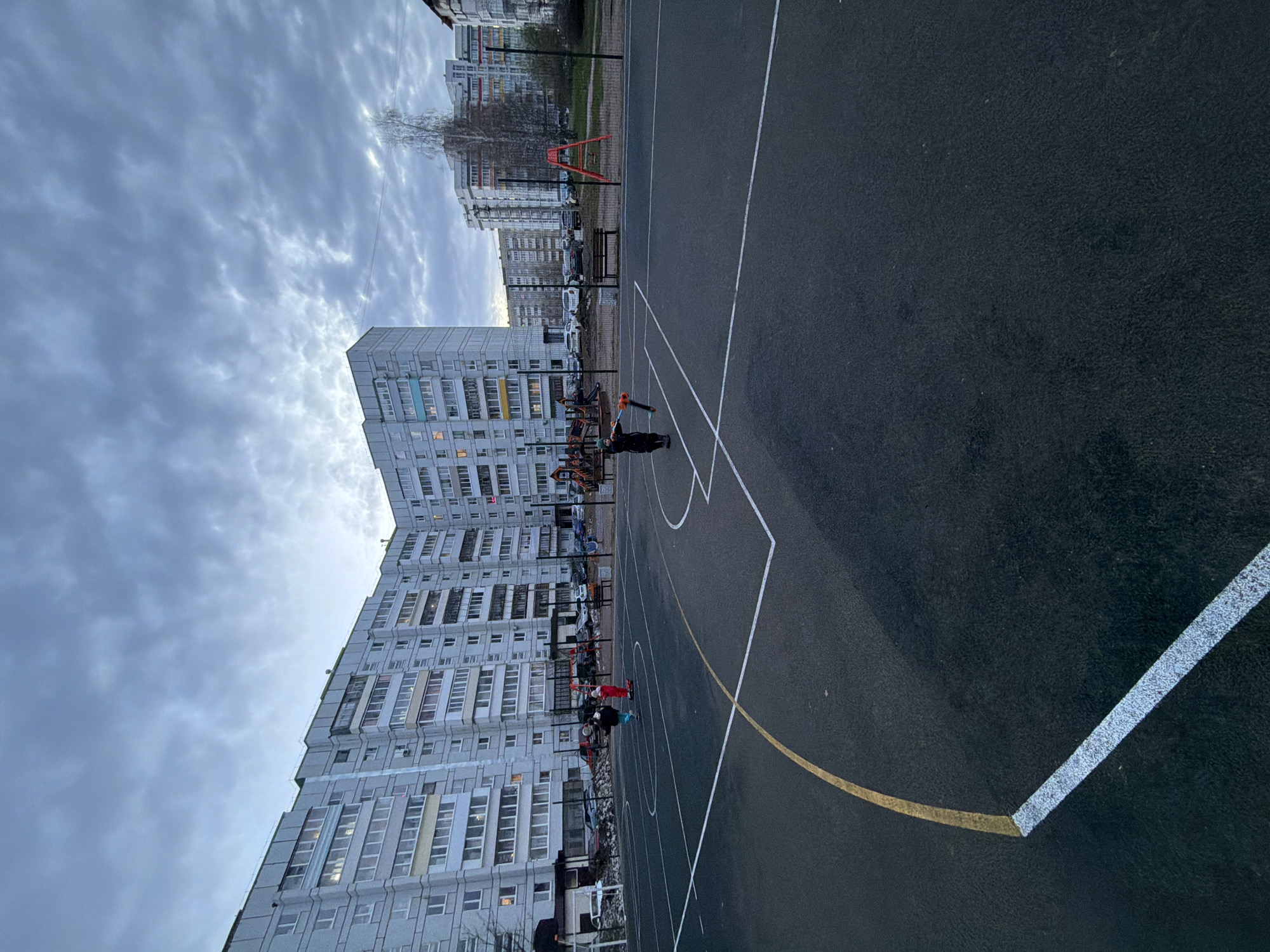 An outdoor scene in Chaikovsky, Russia, depicts a multi-sport court with two individuals on it, set against a backdrop of residential buildings under an overcast sky. The dark grey court is marked with white and yellow lines, suggesting it is used for games like basketball. One person, dressed in dark attire, holds a long orange and black object, possibly a toy or piece of equipment. Another person is also visible further back on the court.
Dominating the background are several tall, multi-story apartment buildings, characteristic of Soviet-era architecture, with numerous windows. Some windows in the buildings show interior lights, hinting at either a late afternoon/early evening setting or a particularly gloomy day. The sky is heavily clouded, creating a diffused light across the scene. Beyond the court and between the buildings, leafless trees, parked cars, and a playground or outdoor workout area with a red structure and a bench are visible. The overall impression is that of a quiet, everyday urban environment. No discernible text is present in the image.