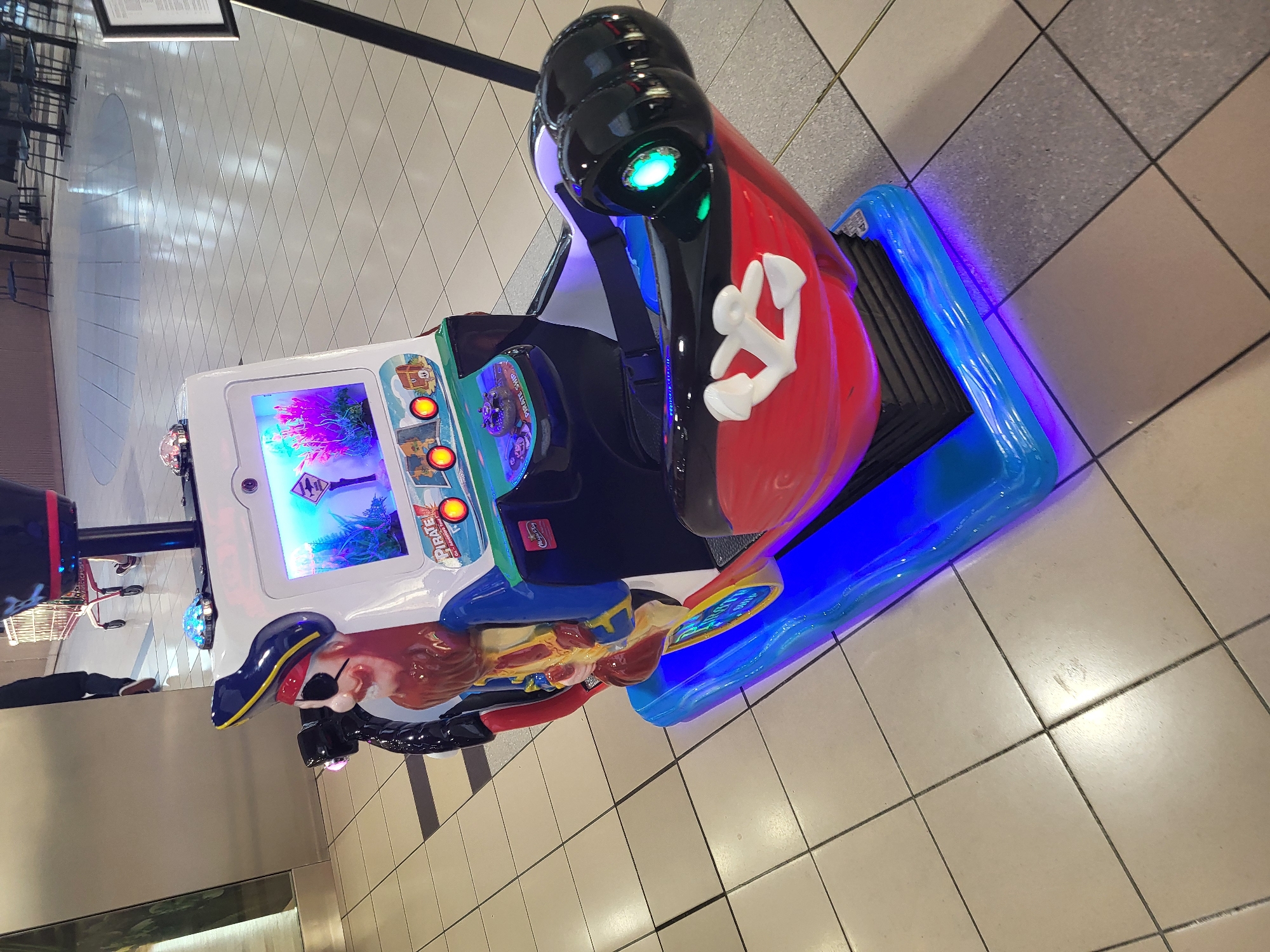 The image shows a pirate-themed ride machine, likely located in a commercial area, possibly a mall in an unknown city. The ride features a screen displaying a pirate-themed video game. The base of the ride is illuminated with blue lights. The flooring consists of light-colored square tiles.