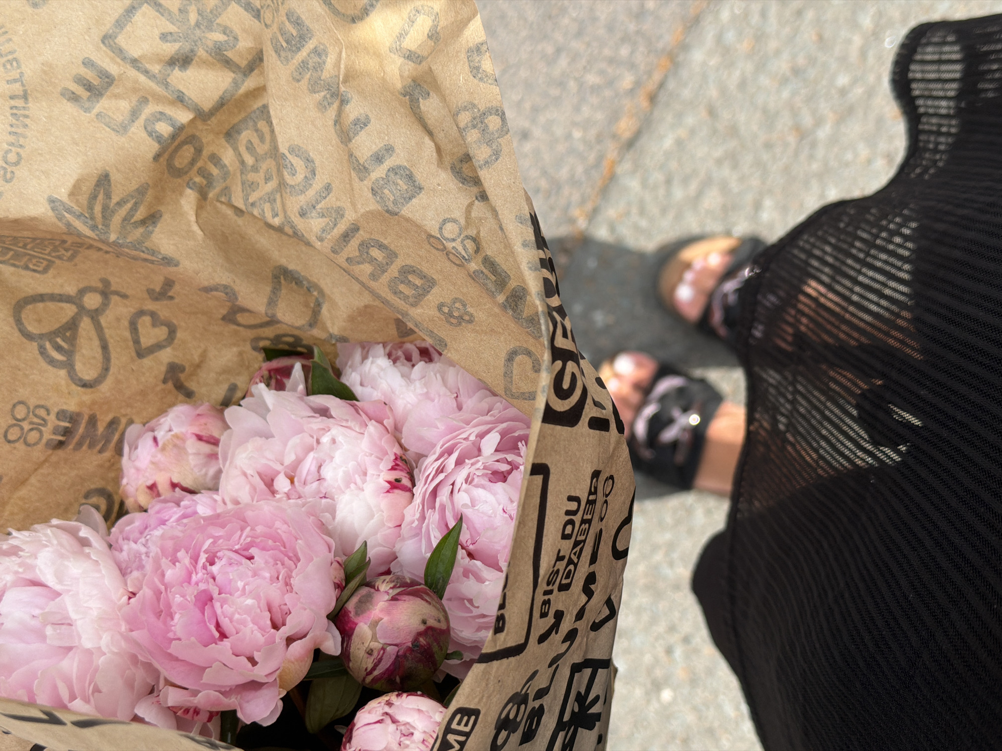 A bouquet of pink peonies is presented in brown paper packaging. The paper is adorned with printed text and illustrations, including bees, hearts, and gift boxes. The bouquet is held by a person wearing black sandals and a black mesh garment. The scene appears to be set on an outdoor paved surface, possibly a sidewalk or street in the city of Erfurt, Germany, judging by the text on the paper packaging which reads "BLUMEN" (flowers in German) and "BIST DU DABEI" (are you there/joining in). The lighting suggests it is daytime, with shadows cast on the ground.