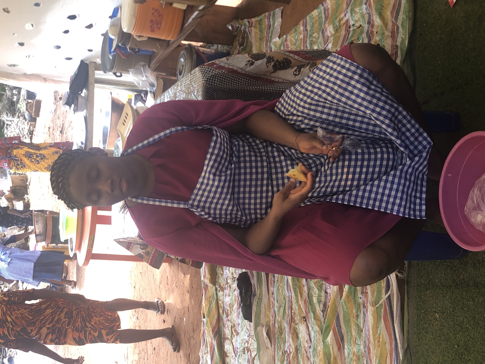 A woman is seated at a table in what appears to be an outdoor market setting. The woman wears a dark pink dress with a blue and white checkered apron. She is holding what looks like a piece of bread in her hands. She is sitting on a blue stool. A pink plastic basin is on the ground next to her. The table is covered with striped fabric. The foreground shows a sandy surface with two partially visible people. A woman is standing beside an orange plastic table with several blue skirted people behind it. Based on visual cues, the location is Nteje, Nigeria.