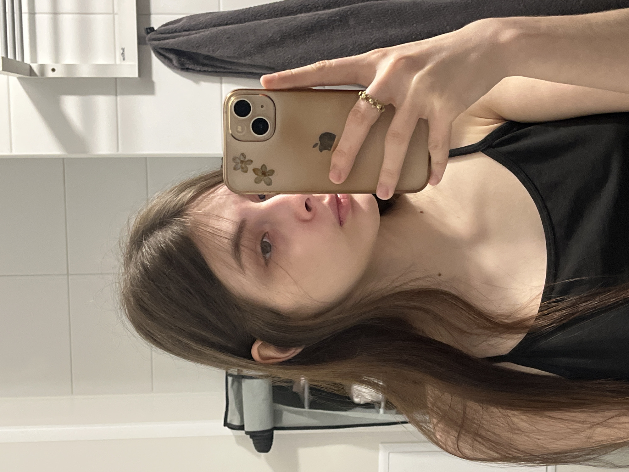 A young woman is taking a selfie in what appears to be a bathroom. She is holding a gold-colored iPhone in front of her face. She has long brown hair and is wearing a black tank top. The background shows white tiled walls and a gray towel rack. There's no indication that this photo was taken in Saint Petersburg, Russia.