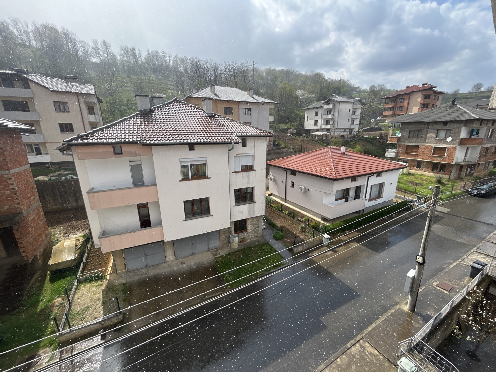 The image depicts a residential neighborhood in Zlatograd, Bulgaria during a period of snowfall. Multiple multi-story buildings with various architectural styles and brick, concrete, and painted facades are visible. Most have pitched roofs. The street is wet, reflecting the ambient light and the ongoing snowfall. Power lines are strung across the street. A hill covered with deciduous trees is in the background. The sky is overcast with low clouds.